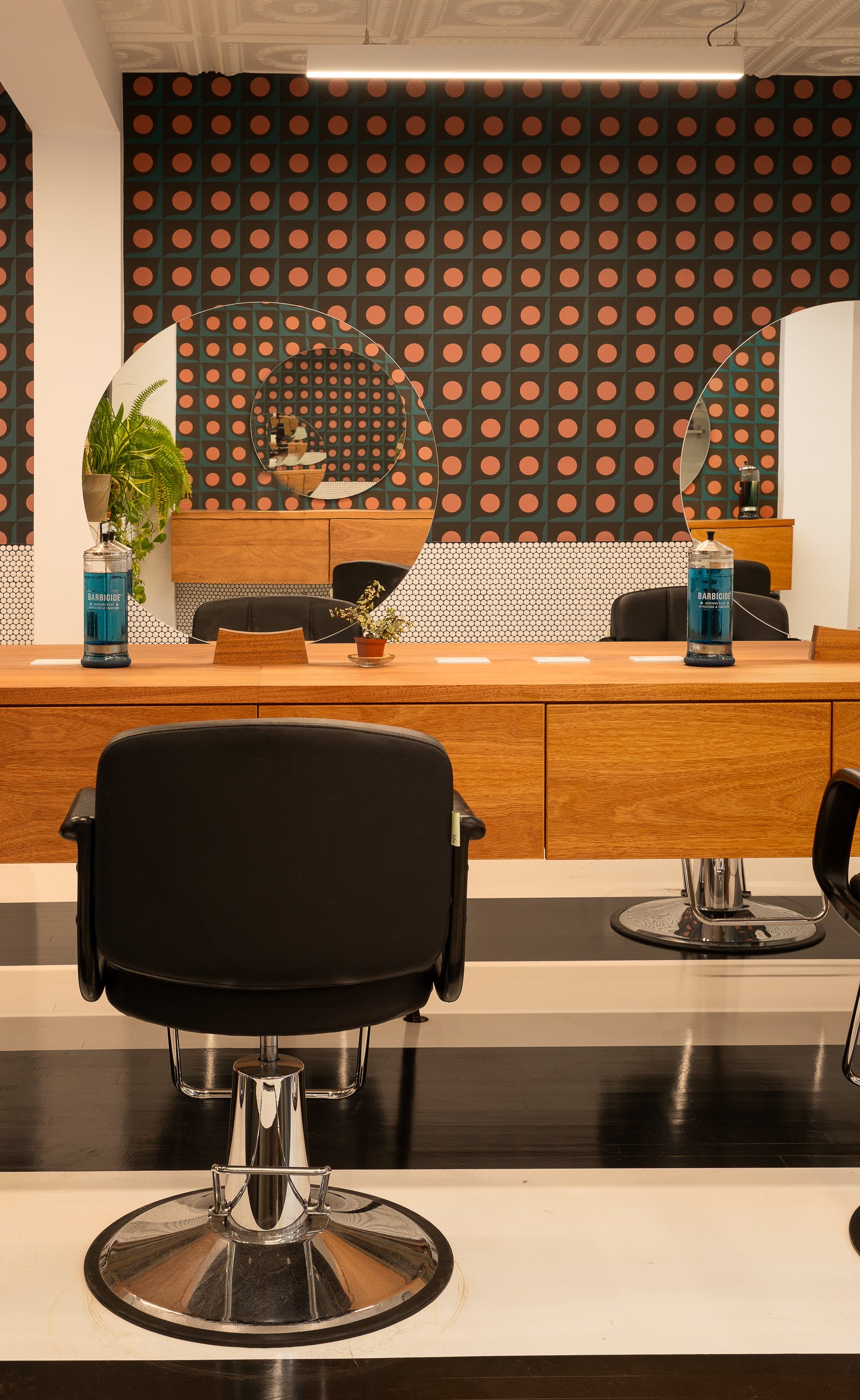 Black salon chair positioned in front of a custom wood styling station with circular mirrors and bold geometric wallpaper.