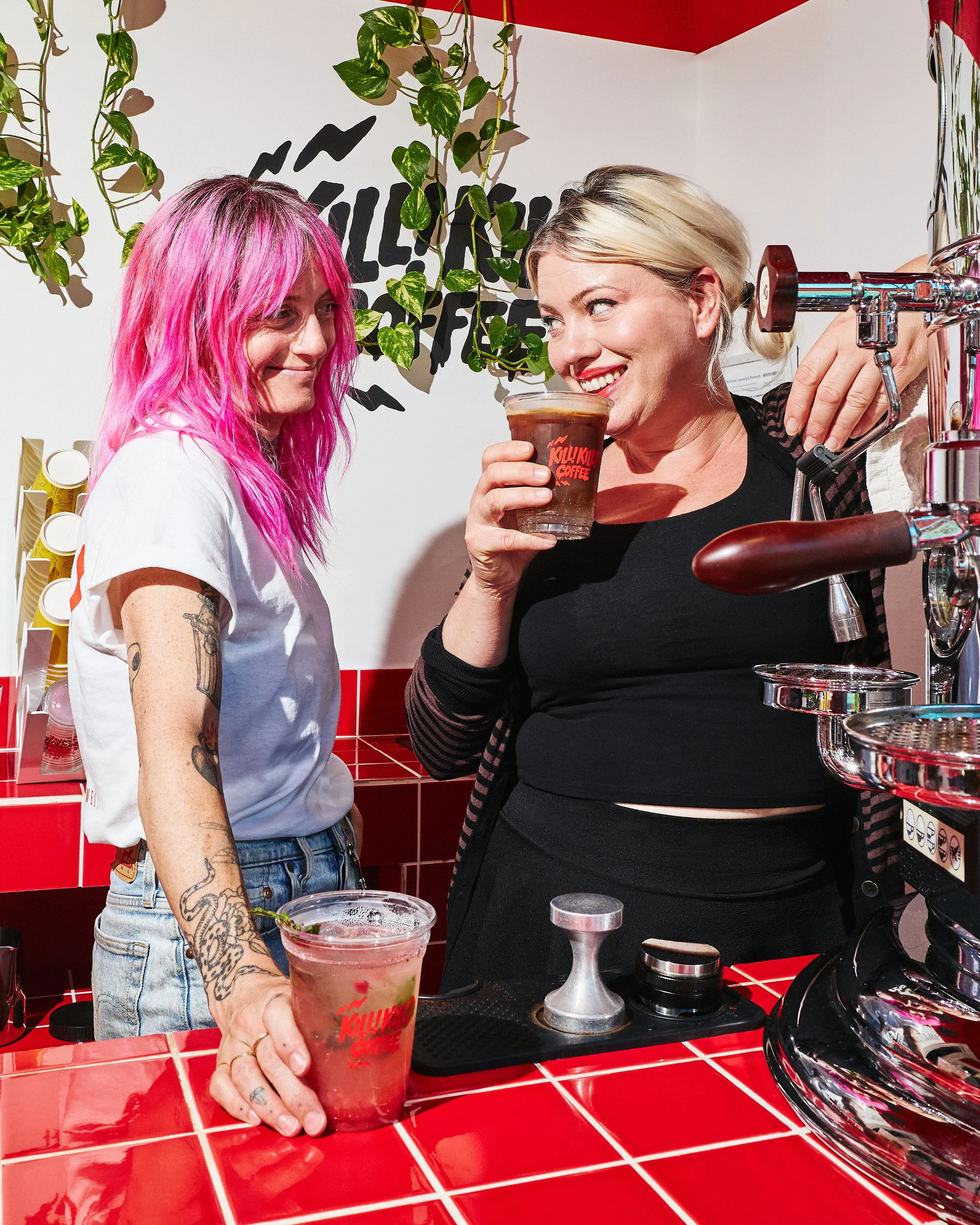 Two people behind the Kill! Kill! Coffee counter with branded iced drinks, red tiles and a chrome espresso machine.