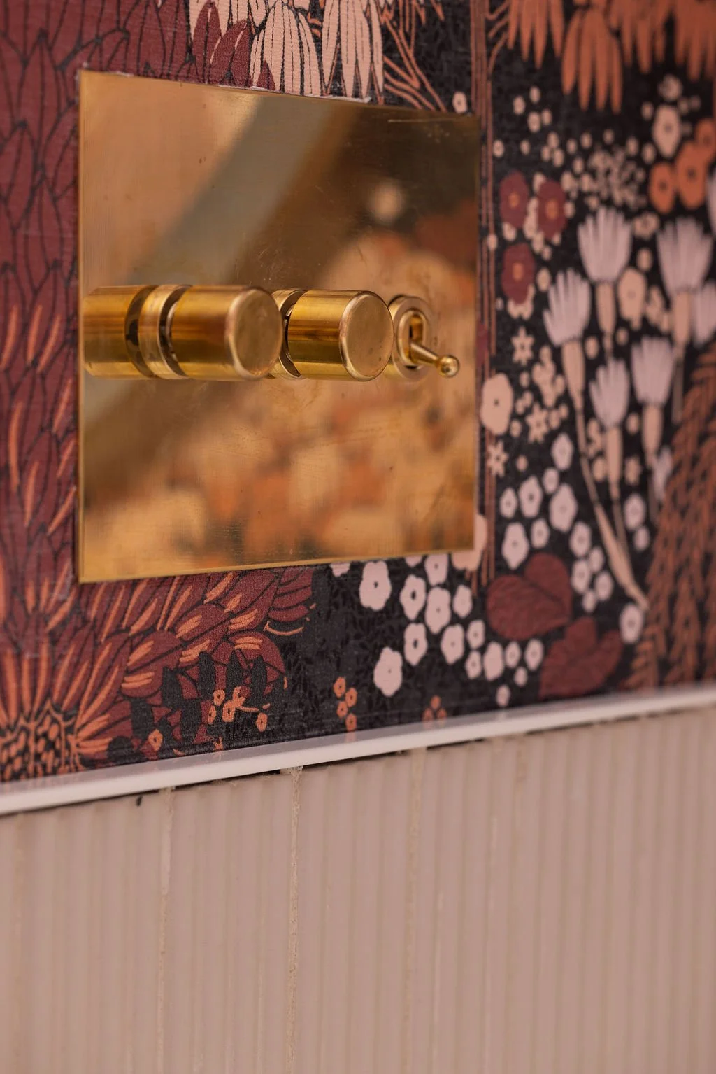 Close-up of brass wall-mounted hardware on a square backplate against warm-toned botanical wallpaper.