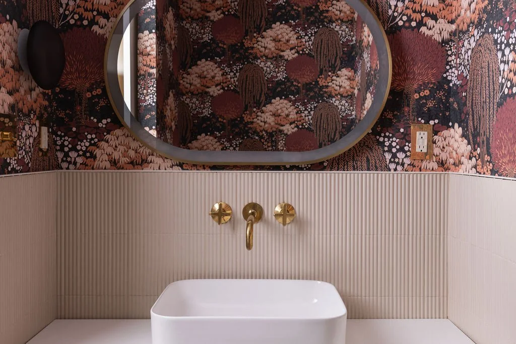 Oval mirror above a white vessel sink and brass wall-mounted faucet, with patterned botanical wallpaper and fluted tile wainscoting.