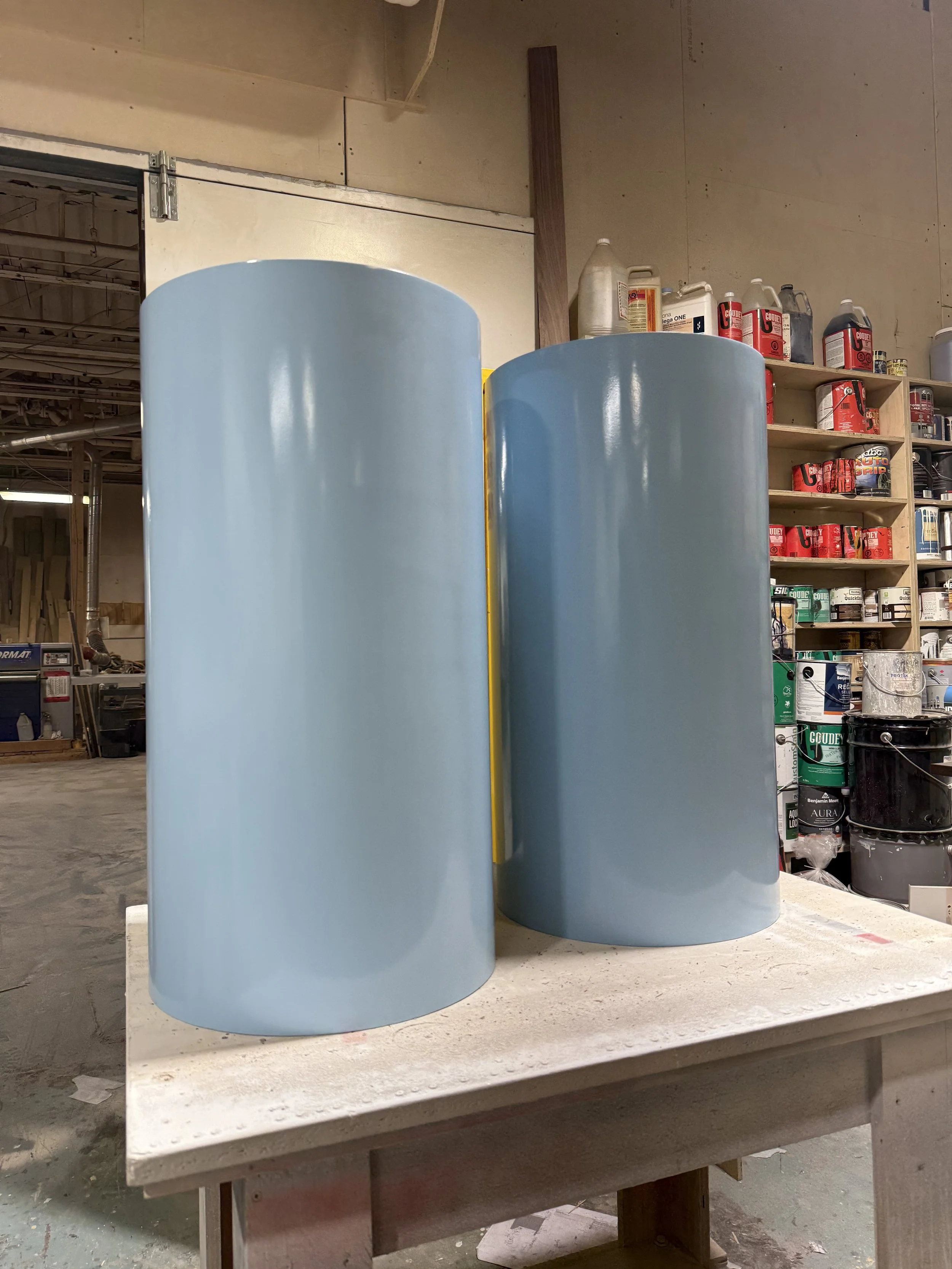 Two pale blue cylindrical table supports with a smooth painted finish during fabrication at the furniture workshop.