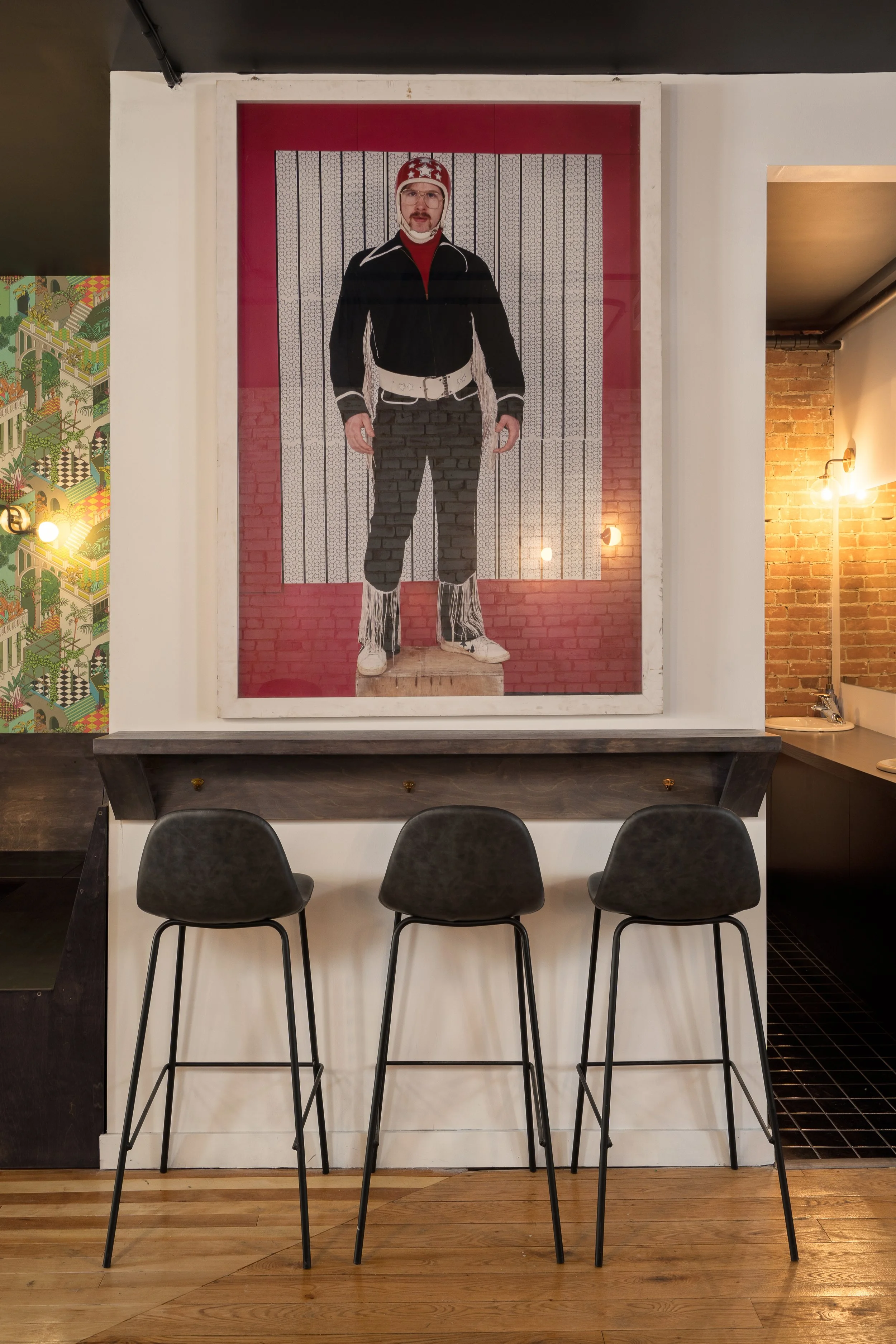 Framed figurative artwork above a dark wood counter with three black stools inside Turbo Haüs.