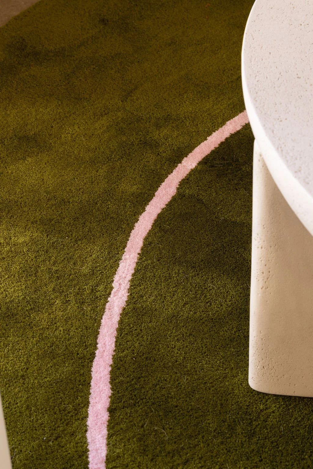 Detail of an olive green area rug with a curved pink line beside a sculptural white coffee table.