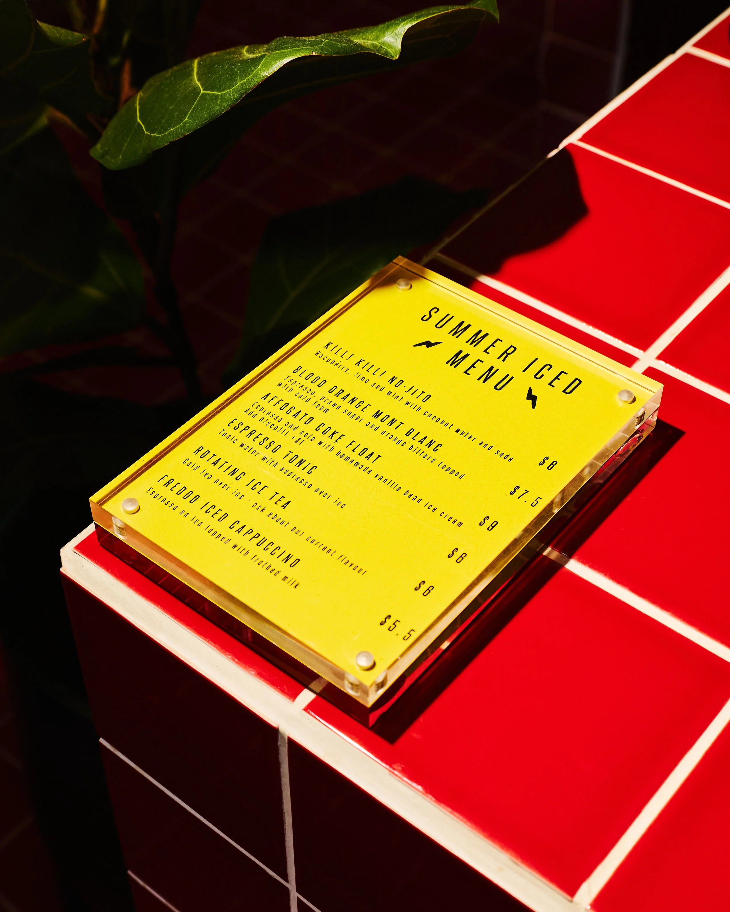 Bright yellow summer iced drinks menu displayed in a clear acrylic holder on the café's red tiled counter.