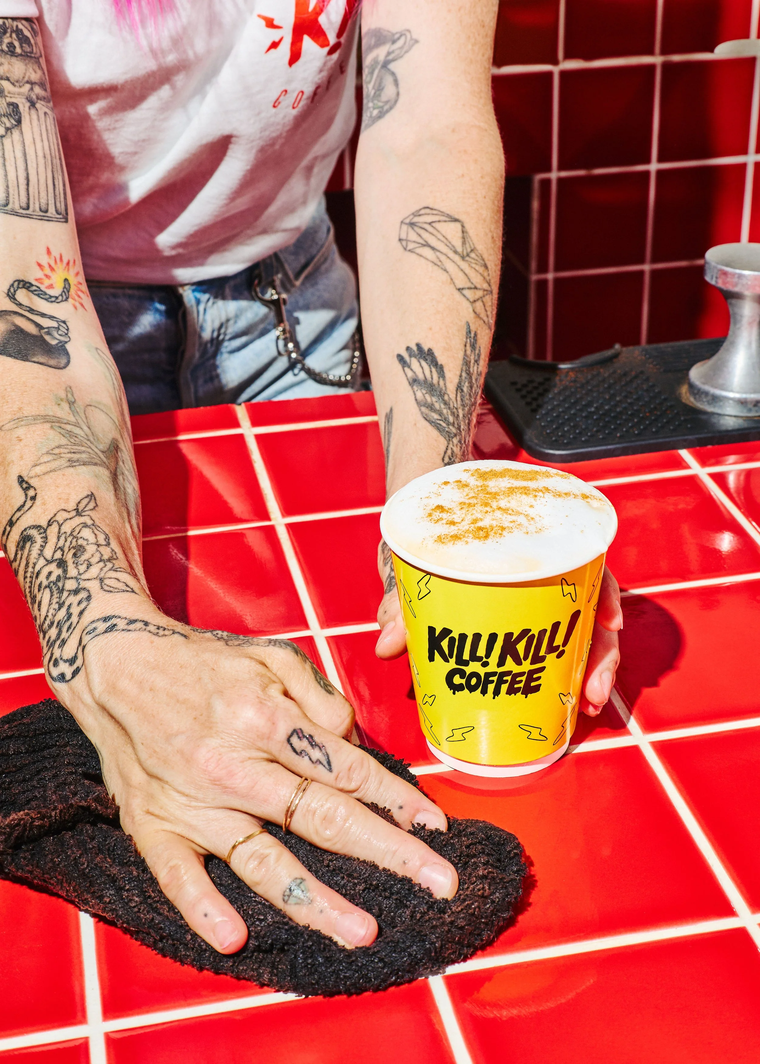 Yellow Kill! Kill! Coffee takeaway cup with black illustrated branding, photographed on the café's red tiled counter.