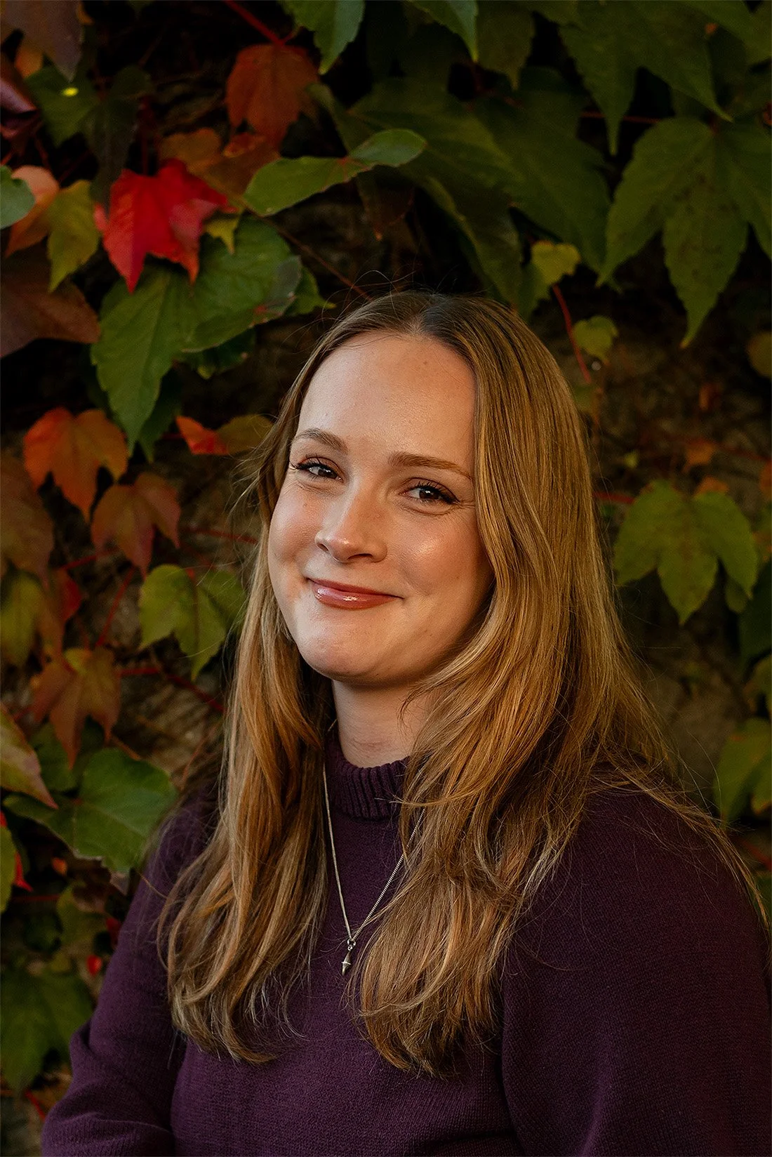 Portrait of a smiling woman with long brown hair wearing a dark sweater in front of autumn foliage