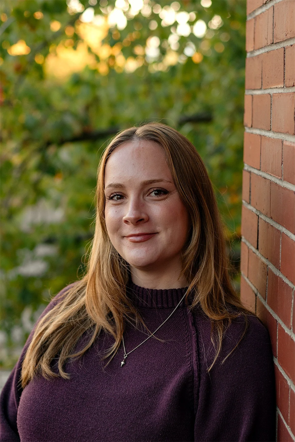 Portrait of a smiling woman in a purple sweater standing beside a brick wall outdoors