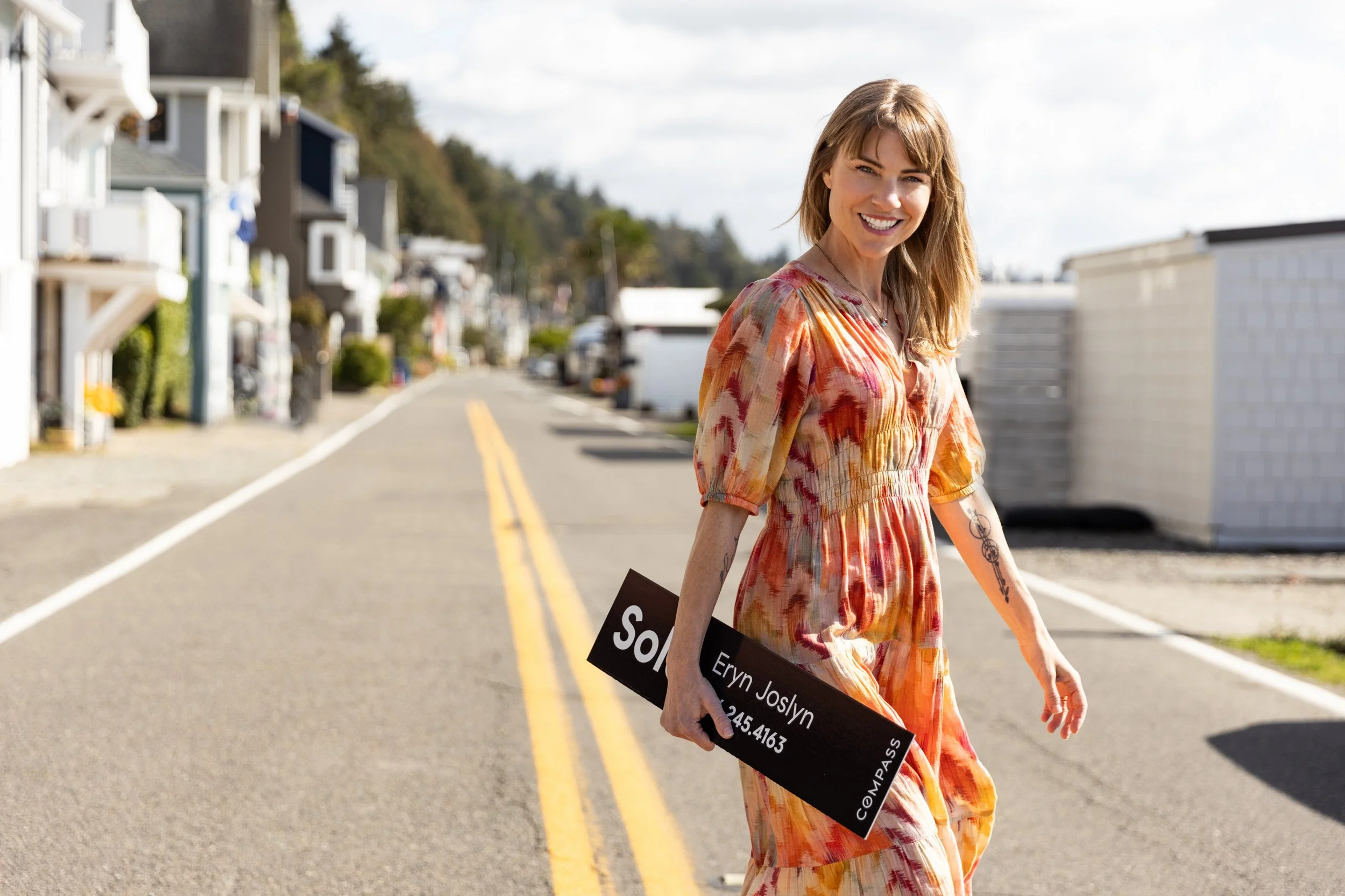 Smiling woman in a patterned dress holding a black casting sign while walking down a residential street