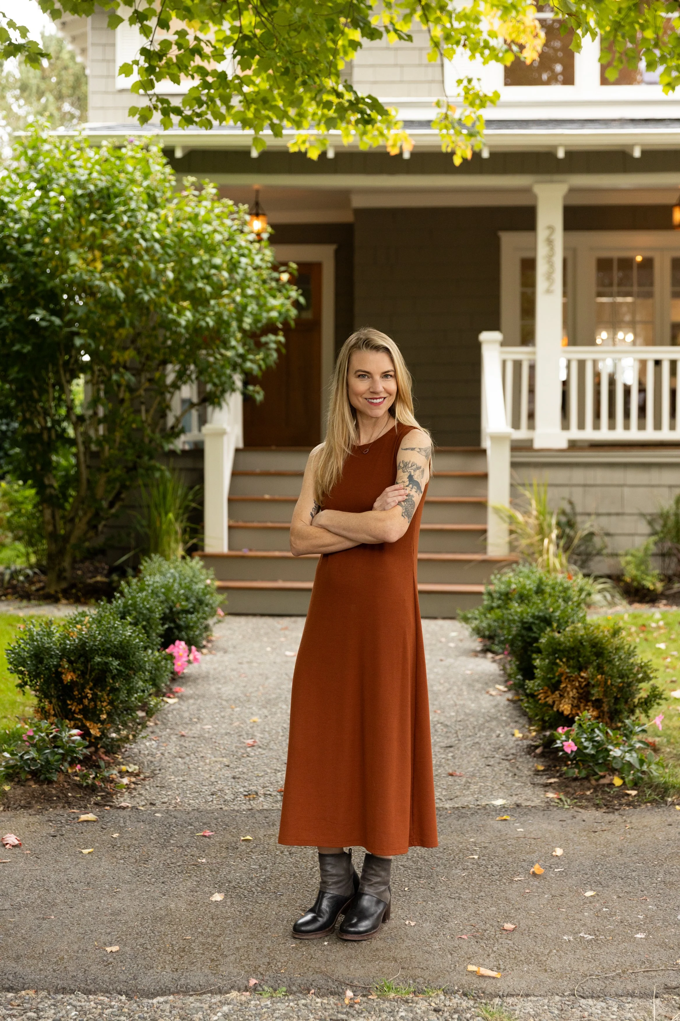 Smiling tattooed woman in a rust-colored dress and black boots standing in a garden outside a house