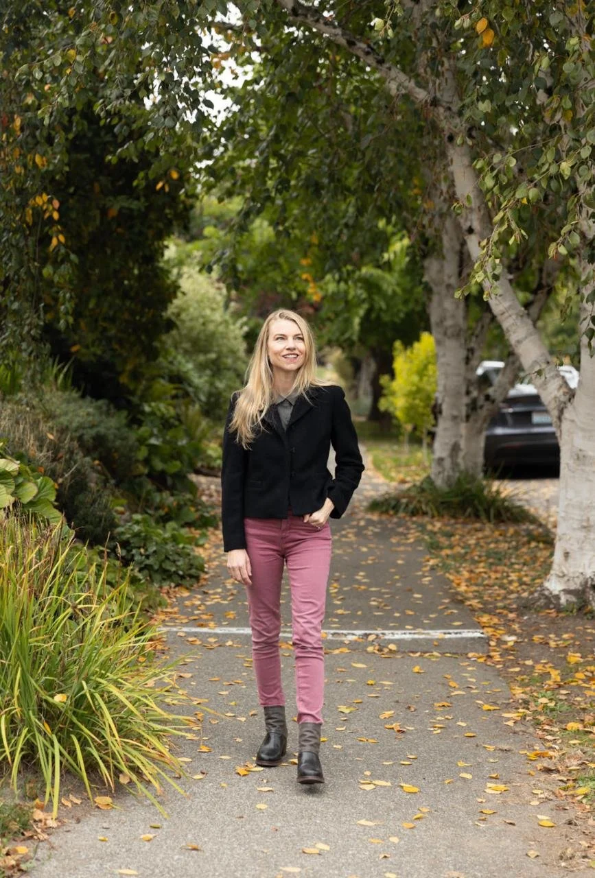 Blonde woman in a black jacket and pink pants walking along a leafy residential sidewalk in autumn