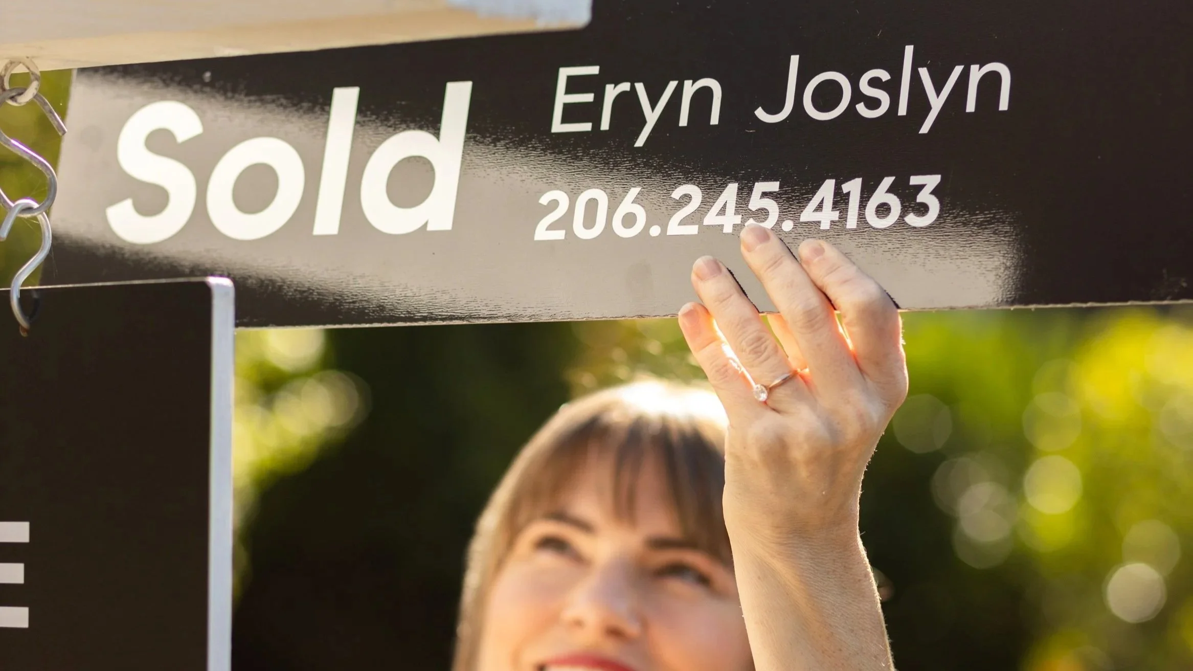 Close-up of a woman holding a black sold sign reading “Eryn Joslyn” and “206.245.4163” outdoors