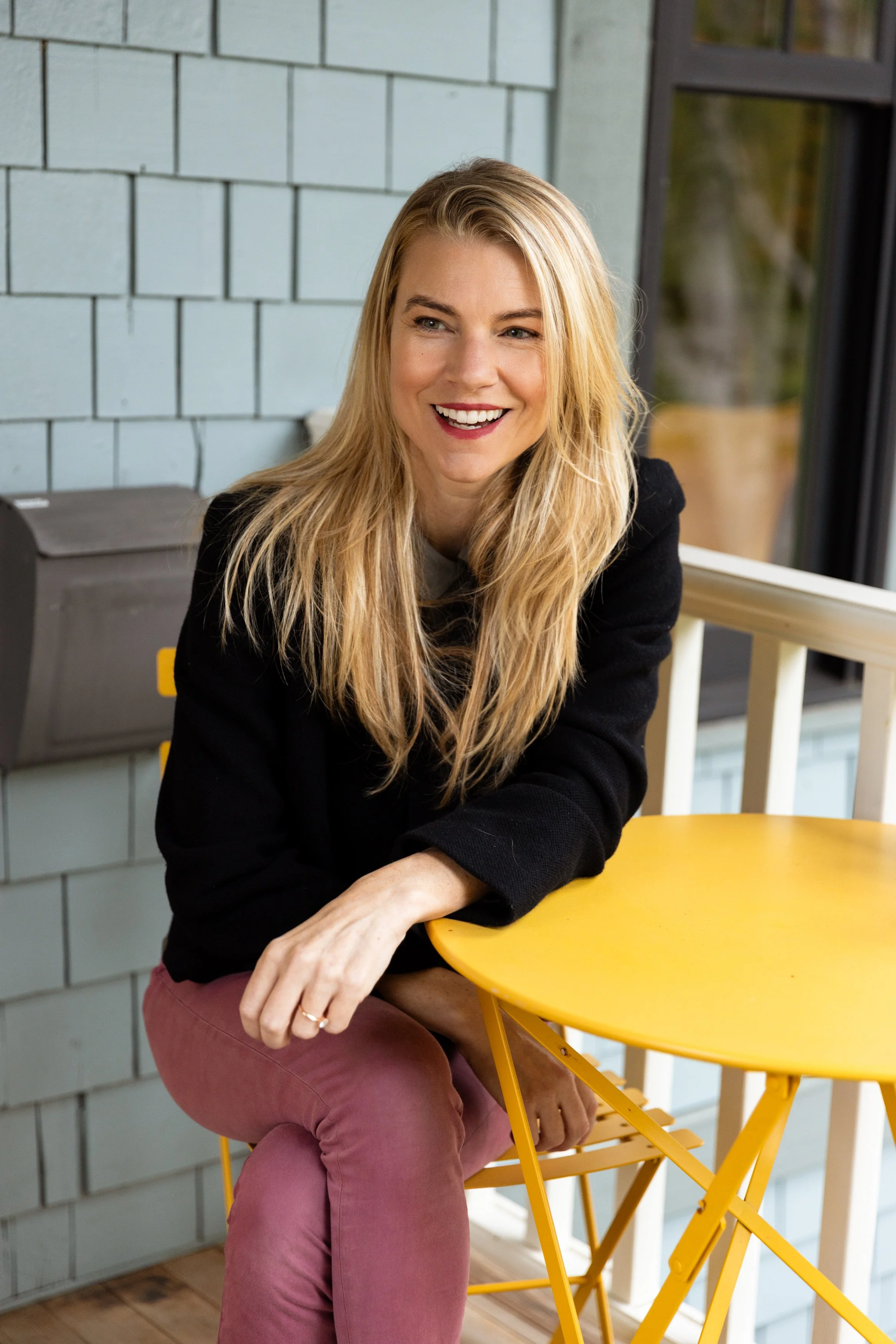 Smiling blonde woman in a black top and burgundy pants sitting at a bright yellow table indoors.