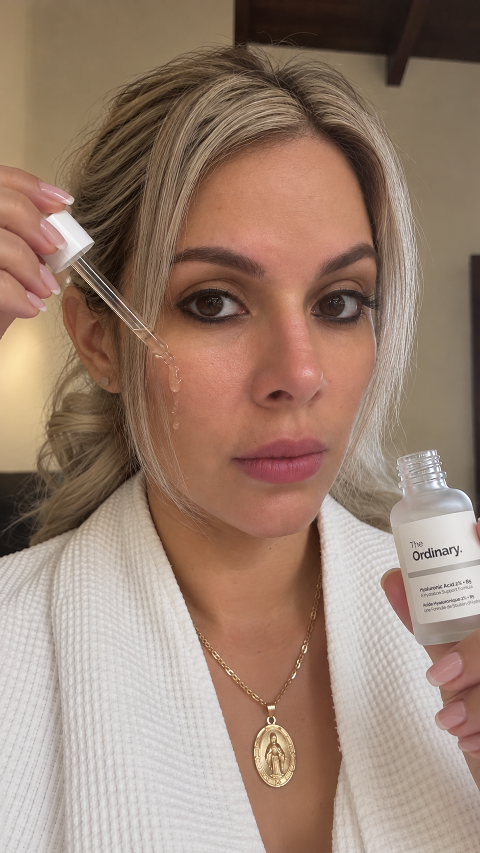 Mujer con cabello rubio y vestido blanco sosteniendo un frasco y una pipeta con sérum en un ambiente interior.