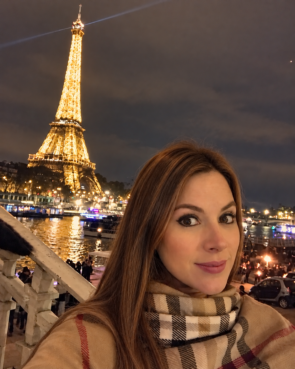 Una mujer con bufanda descubierta en un lugar con vista a la Torre Eiffel iluminada en París, con personas y barcos en el río al fondo y un cielo nocturno.