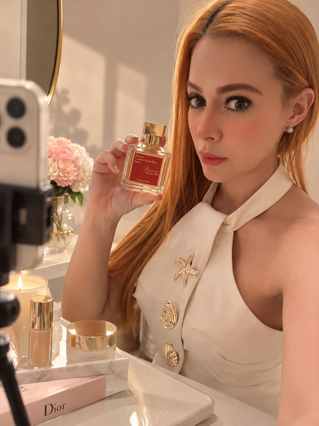 Mujer con cabello rojo y vestido blanco mostrando un perfume Dior en un espejo, con velas y productos de belleza en mesa.