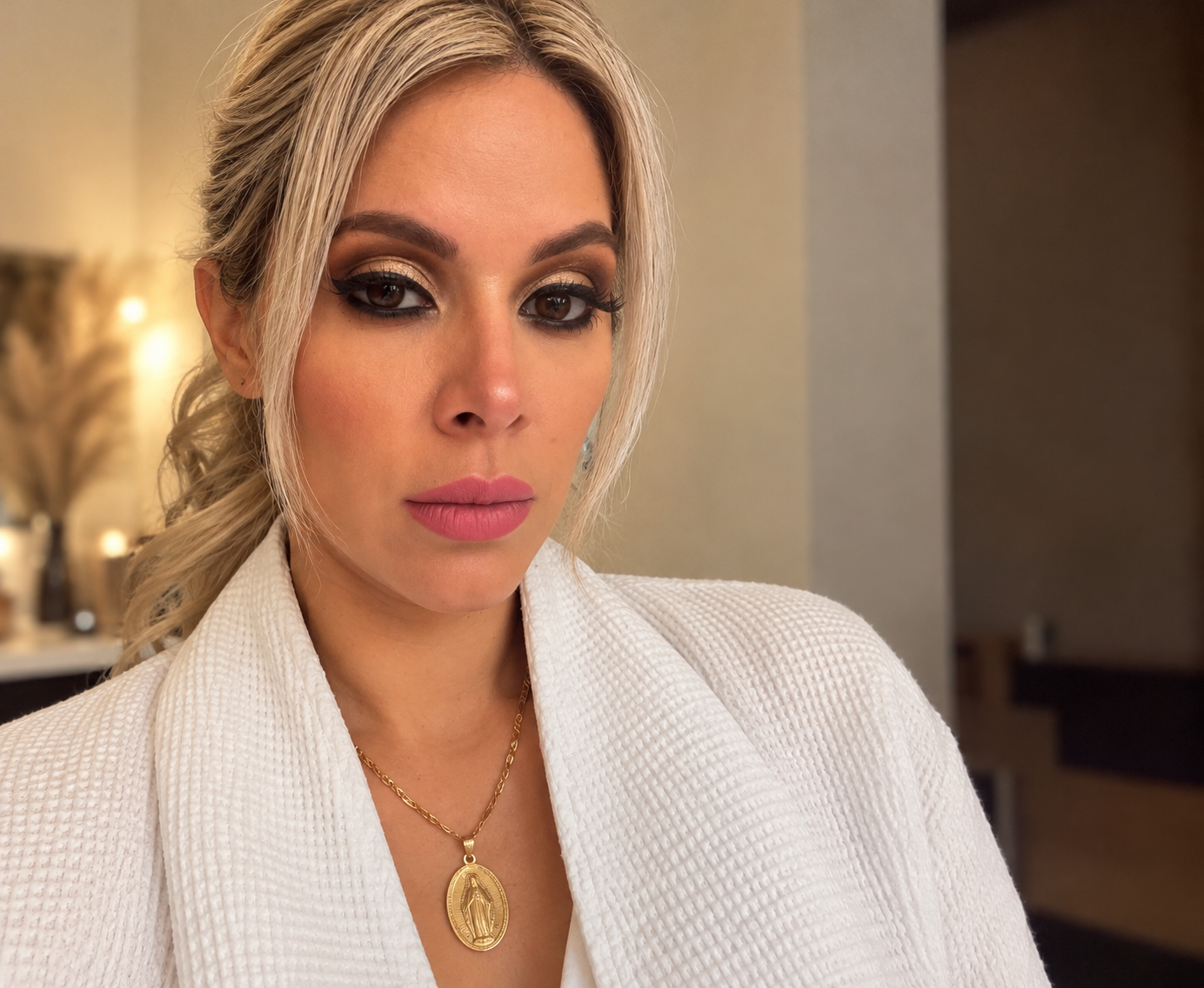 Retrato de una mujer con maquillaje elaborado, vestido blanco y collar con medalla en un entorno interior.