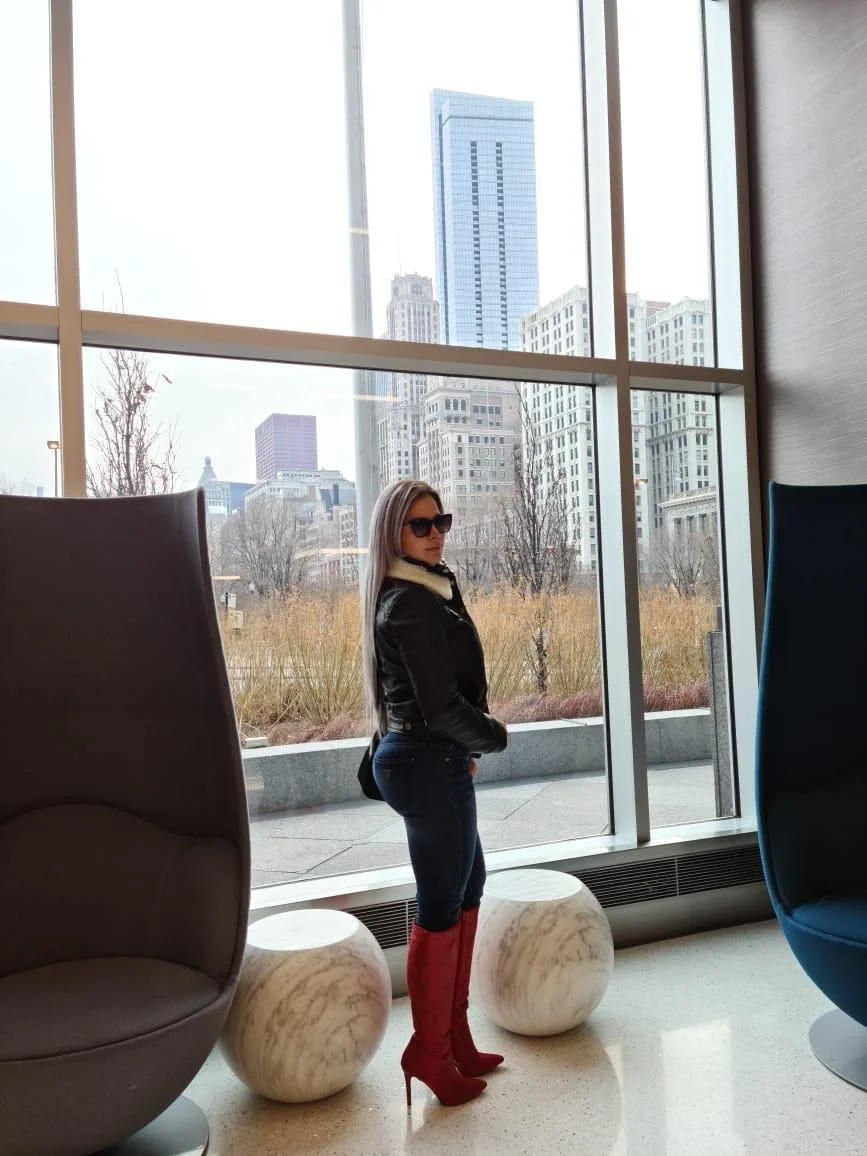 Mujer con gafas oscuras y botas rojas de tacón alto, vestida con chaqueta negra y pantalones ajustados, posando en un interior con ventanas grandes y vista a un centro urbano con edificios altos.