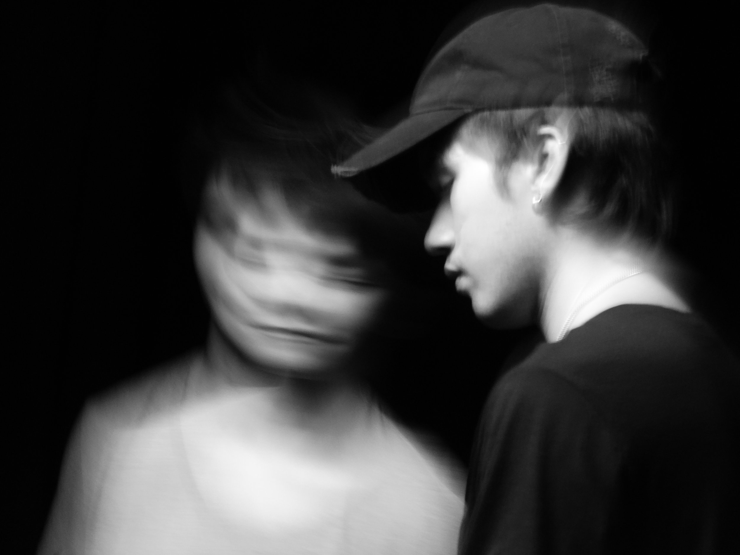 Black-and-white photo of two people in hats embracing closely, with one motion-blurred face.