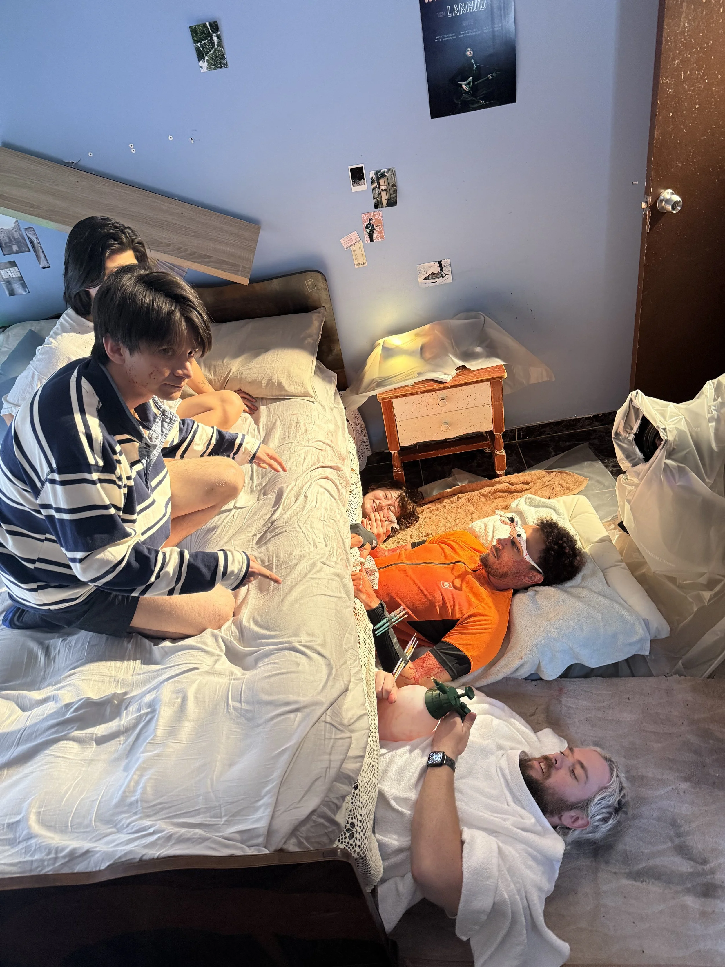 Group of people gathered around beds in a cluttered bedroom with wall photos, blankets, and a green medical device.