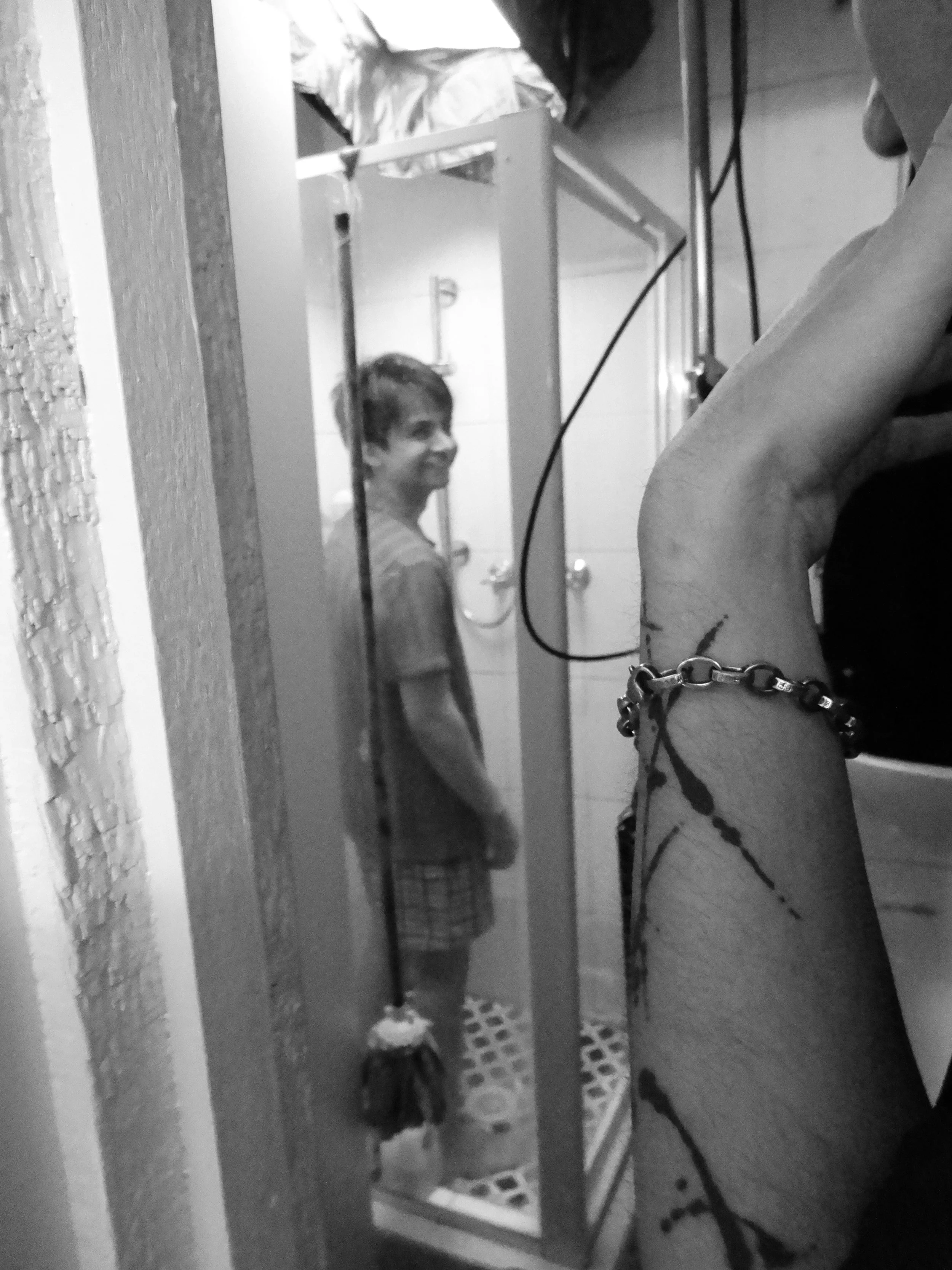 Black-and-white photo of a smiling person standing in a doorway behind another person wearing glasses and a beaded necklace.