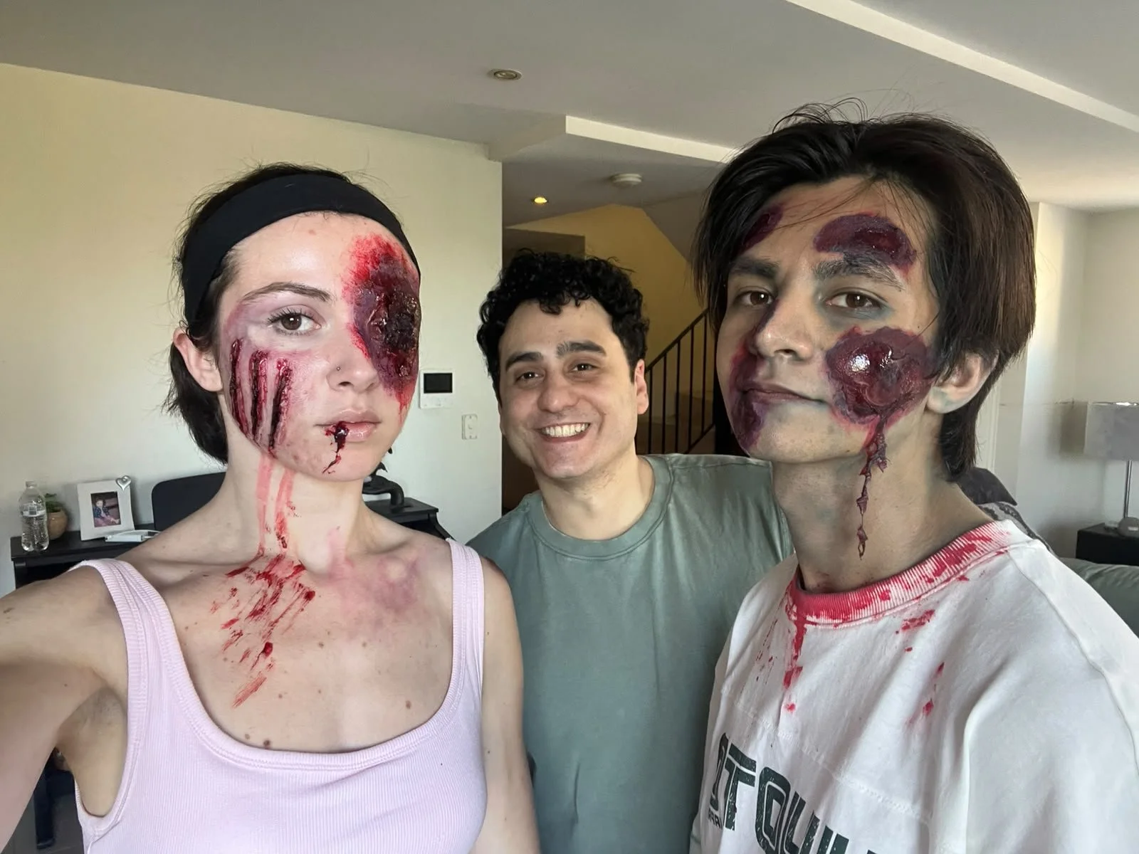 Three people pose indoors, with two wearing realistic zombie makeup featuring blood, bruises, and facial wounds.