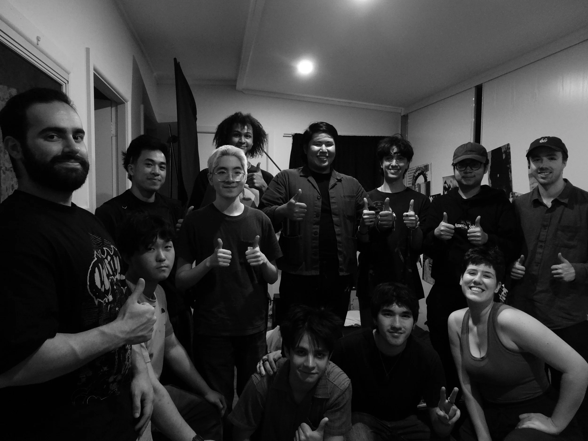 Black-and-white group portrait of 13 people posing indoors with thumbs-up gestures.