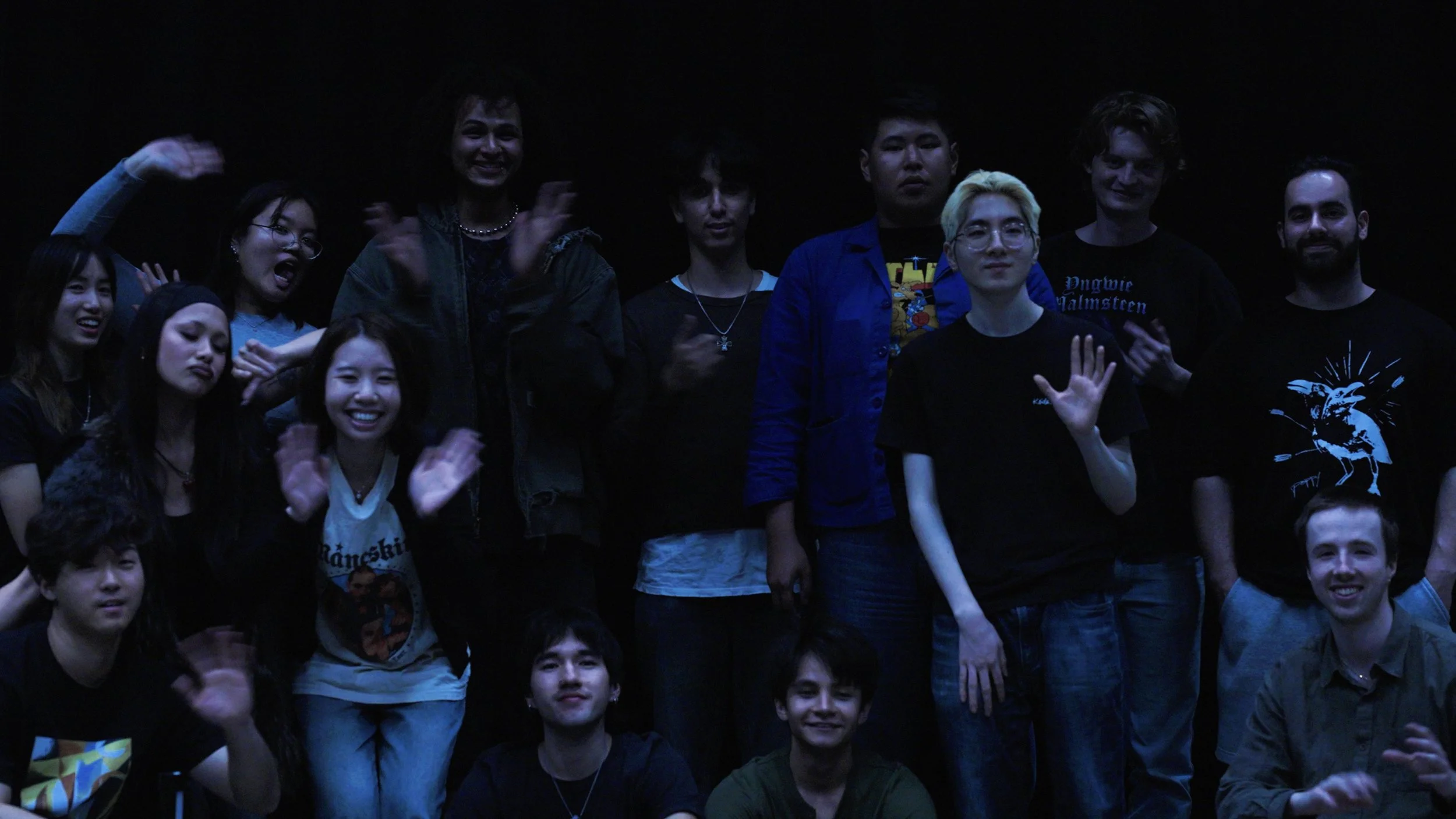 Large group of people posing and waving together in a dimly lit room.
