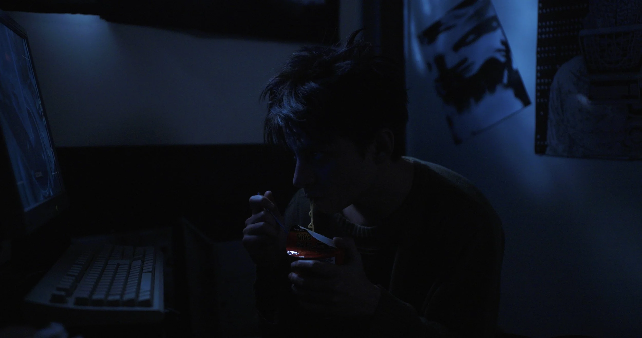 Person eating noodles in a dimly lit room beside a computer monitor