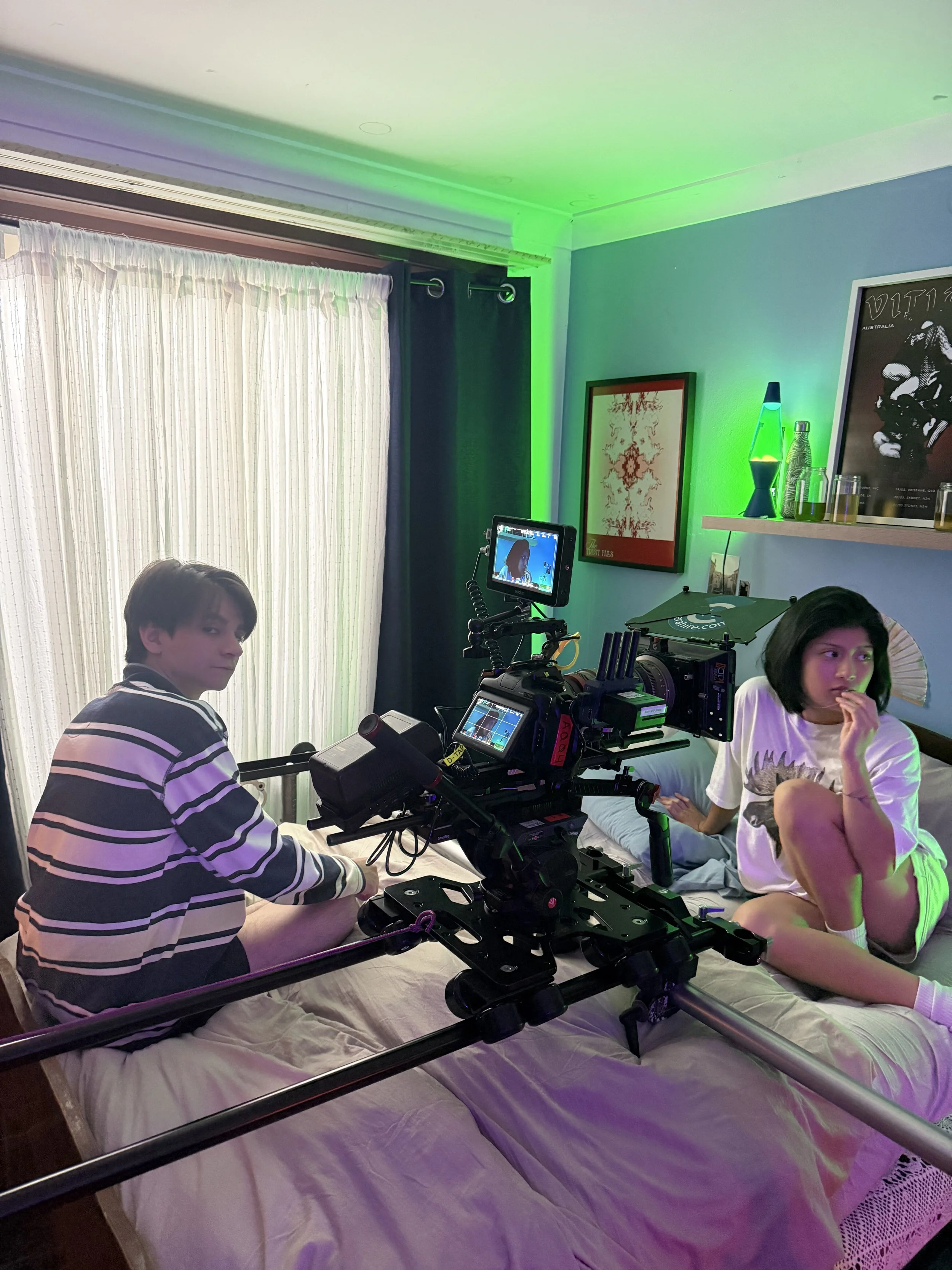 Two people sit on a bed beside a professional cinema camera rig in a bedroom lit with green and purple lights.