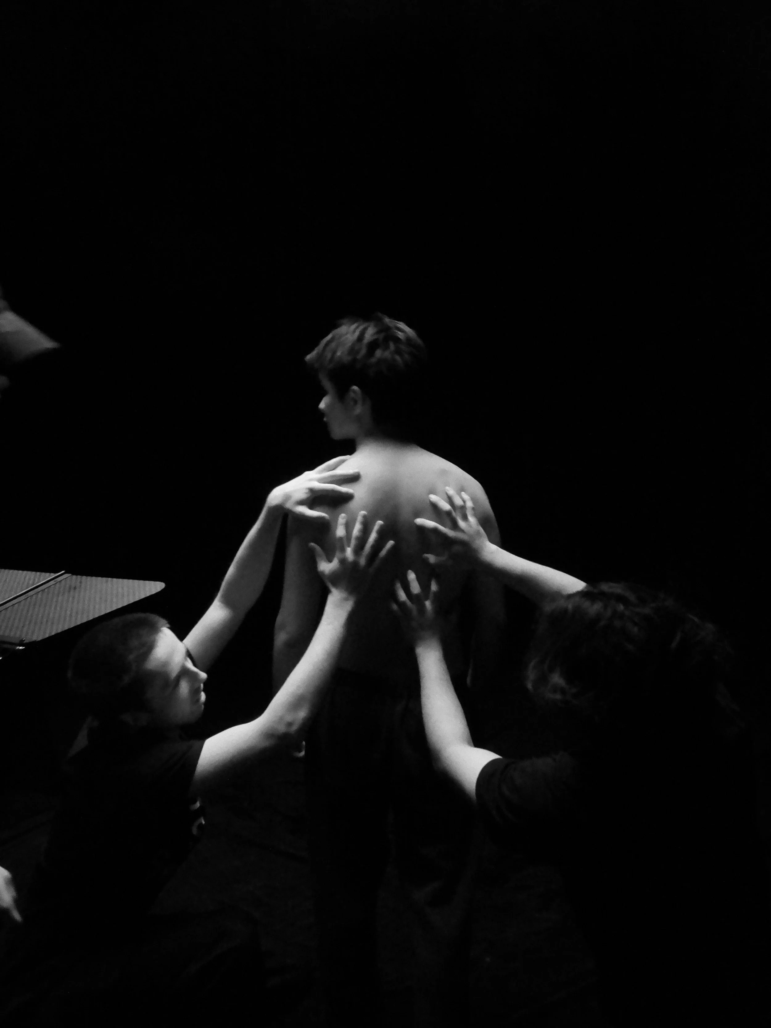 Black-and-white performance photo of a shirtless person surrounded by multiple reaching hands against a dark background