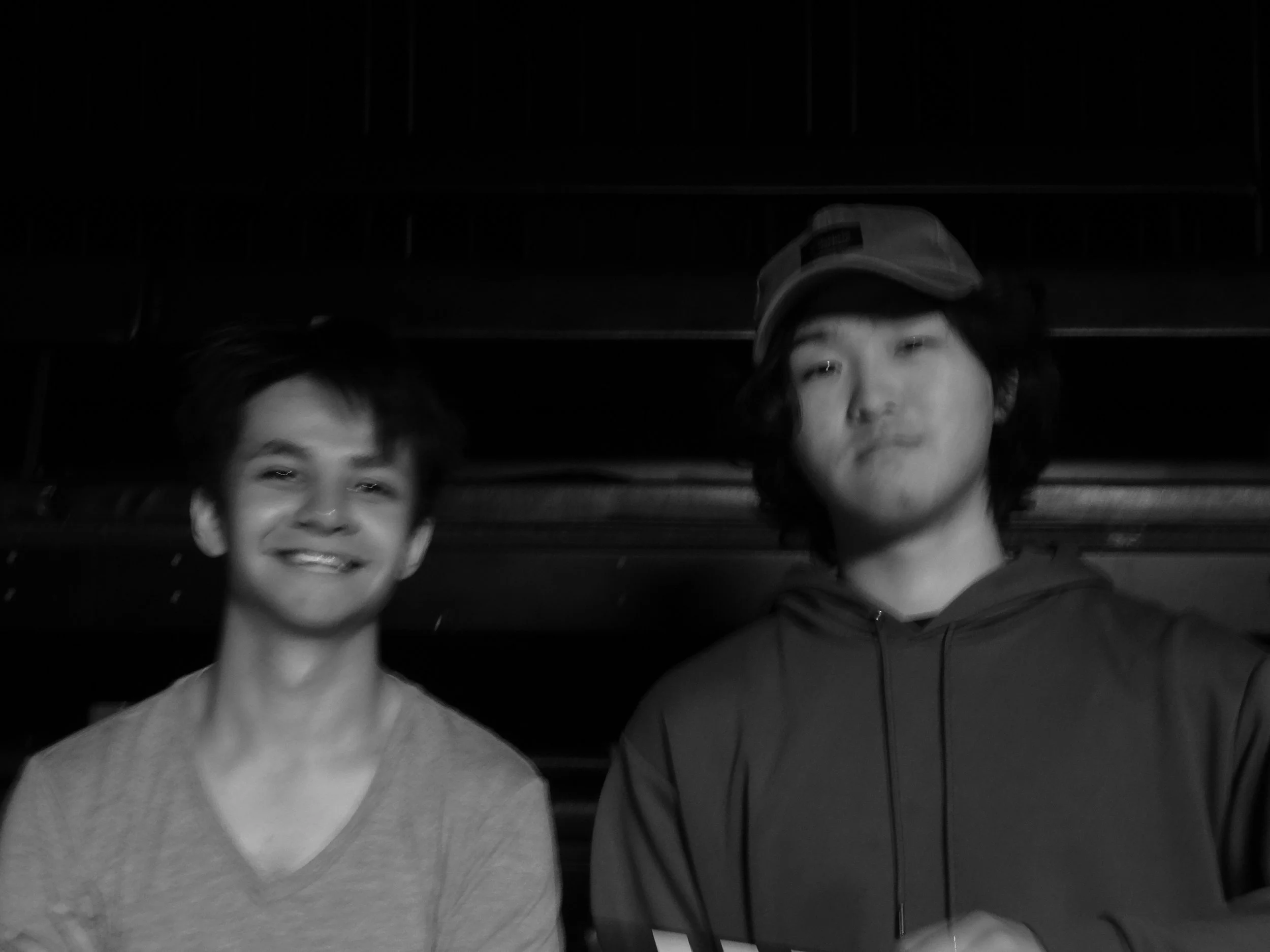 Black-and-white photo of two people smiling and posing together indoors, with one wearing a baseball cap and hoodie.