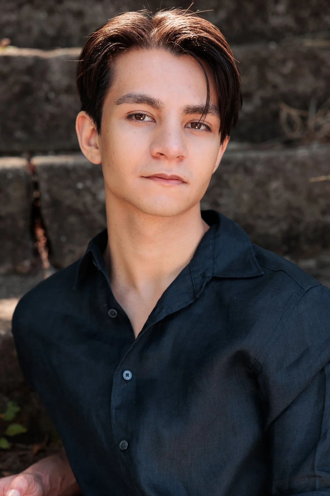A young man with brown hair and light skin wearing a black button-up shirt, standing outdoors with a blurred rocky background.