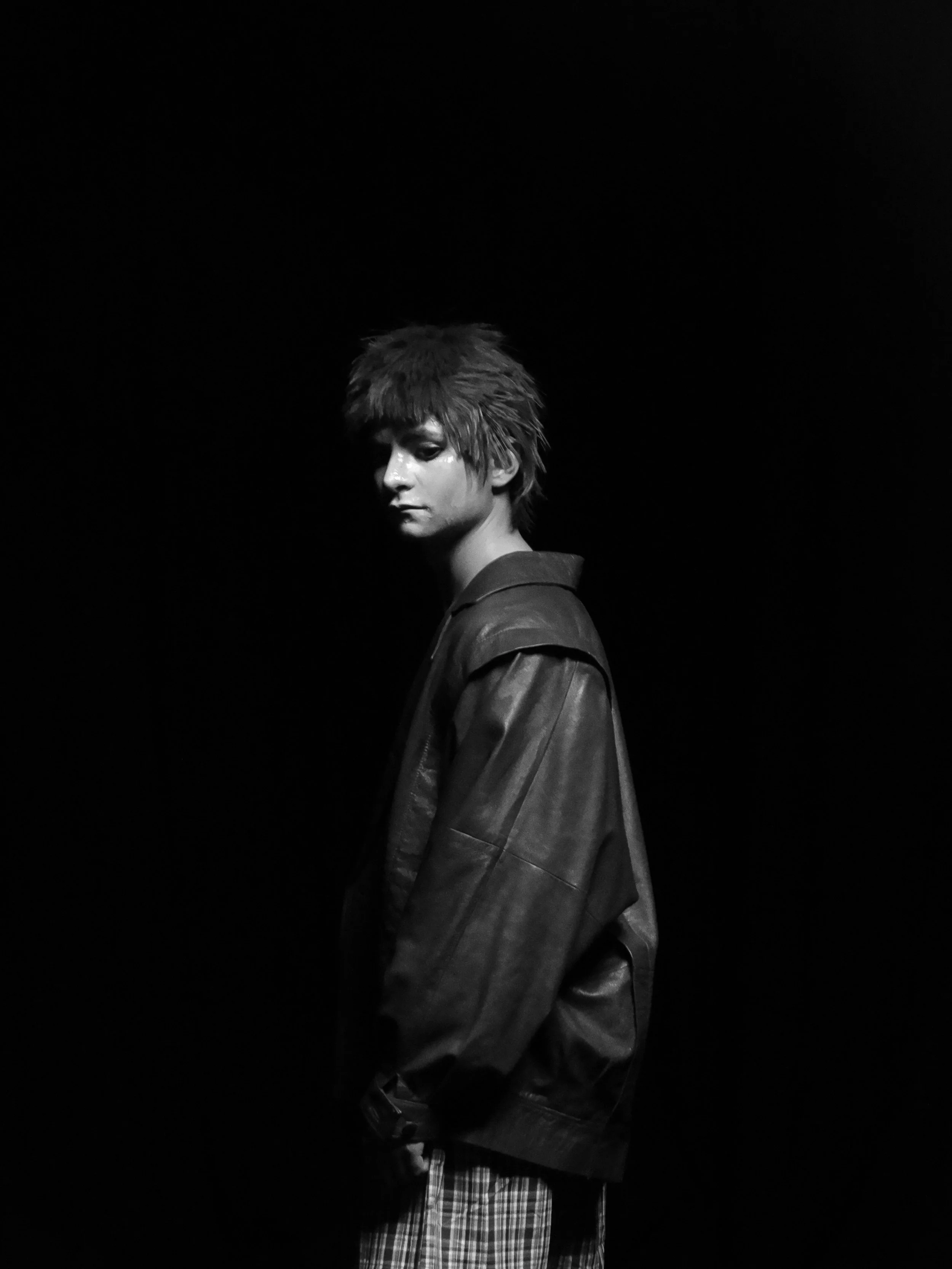 Black-and-white portrait of a person with shaggy hair wearing a dark jacket against a black background.