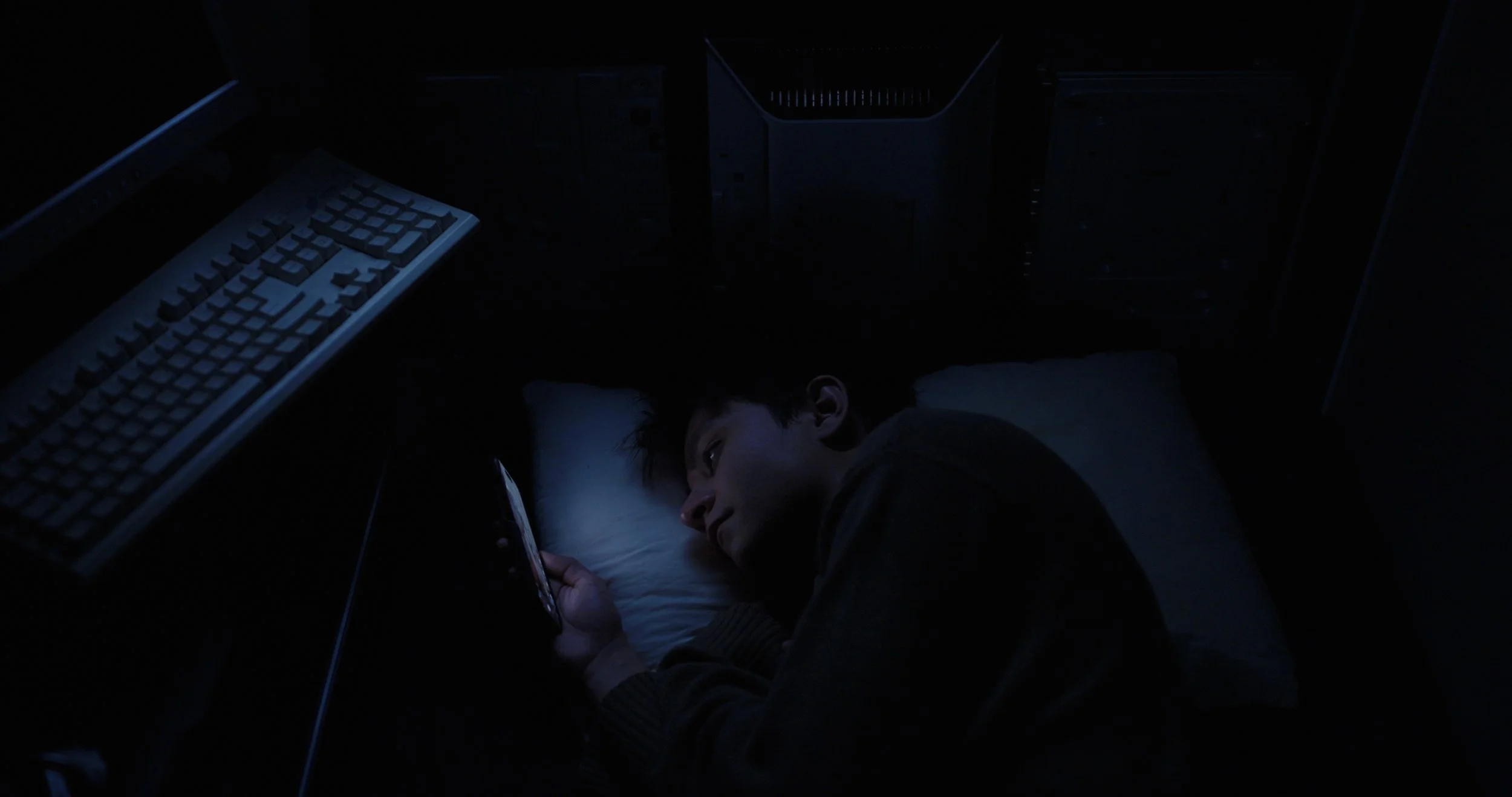 Person lying in bed at night using a smartphone beside a desktop computer keyboard