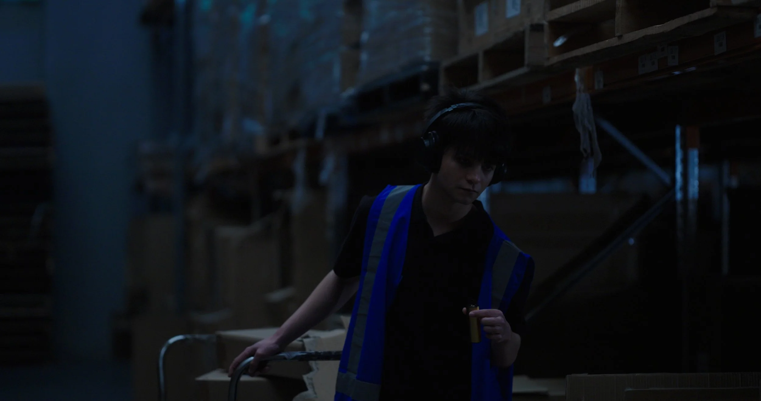 Warehouse worker wearing a reflective safety vest and headphones moving a cart among stacked boxes in a dim storage area