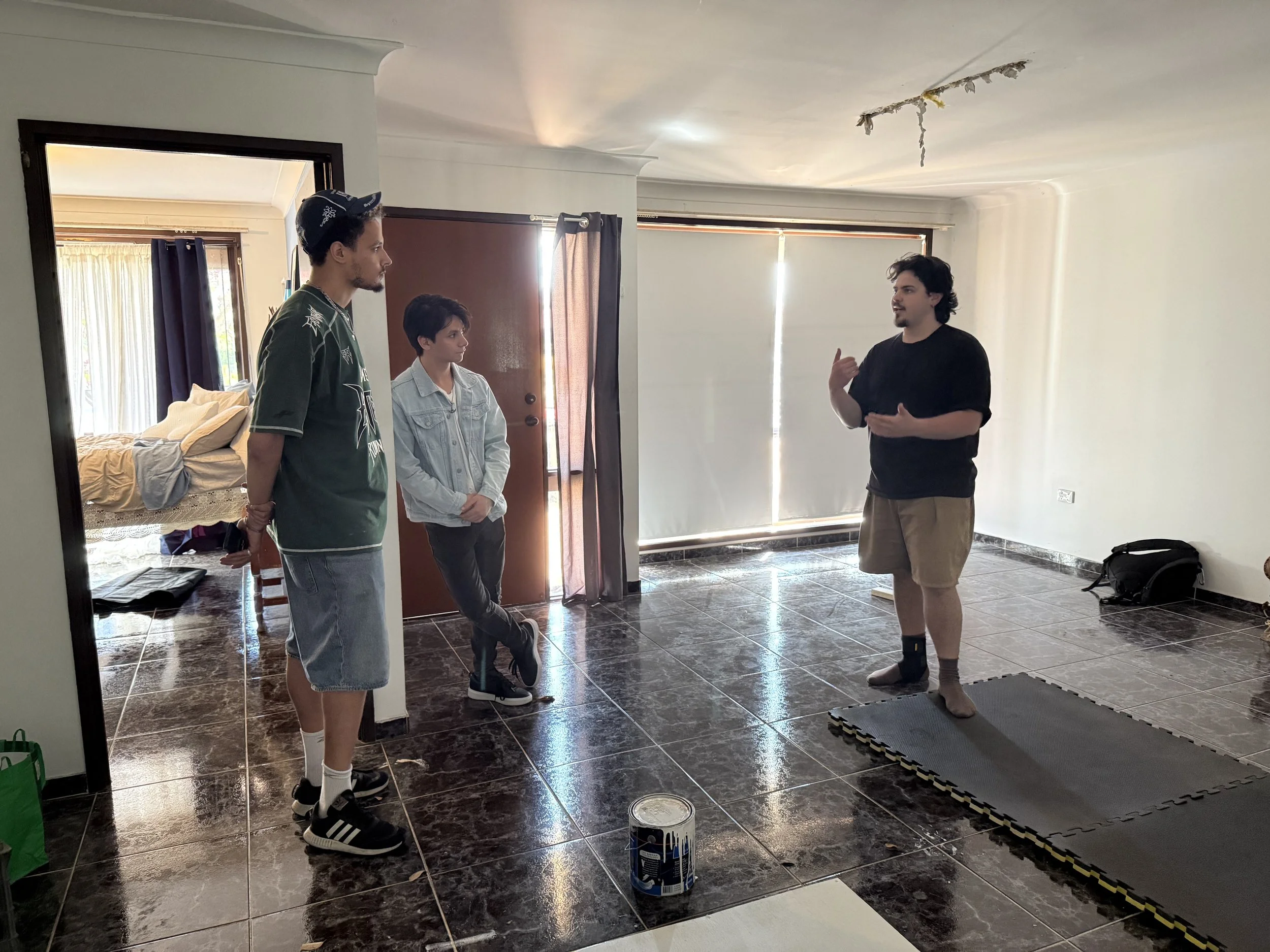 Three men talk in a partially renovated room with tiled floors, paint cans, and exercise mats.
