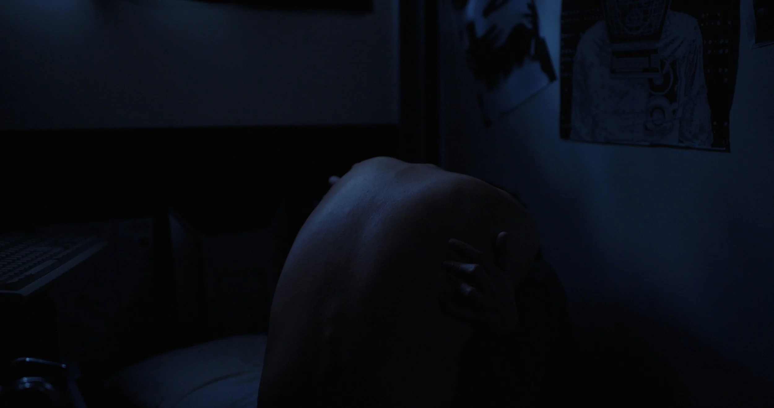 Dark blue-lit bedroom with a person curled up under a blanket, framed by a keyboard and wall artwork.