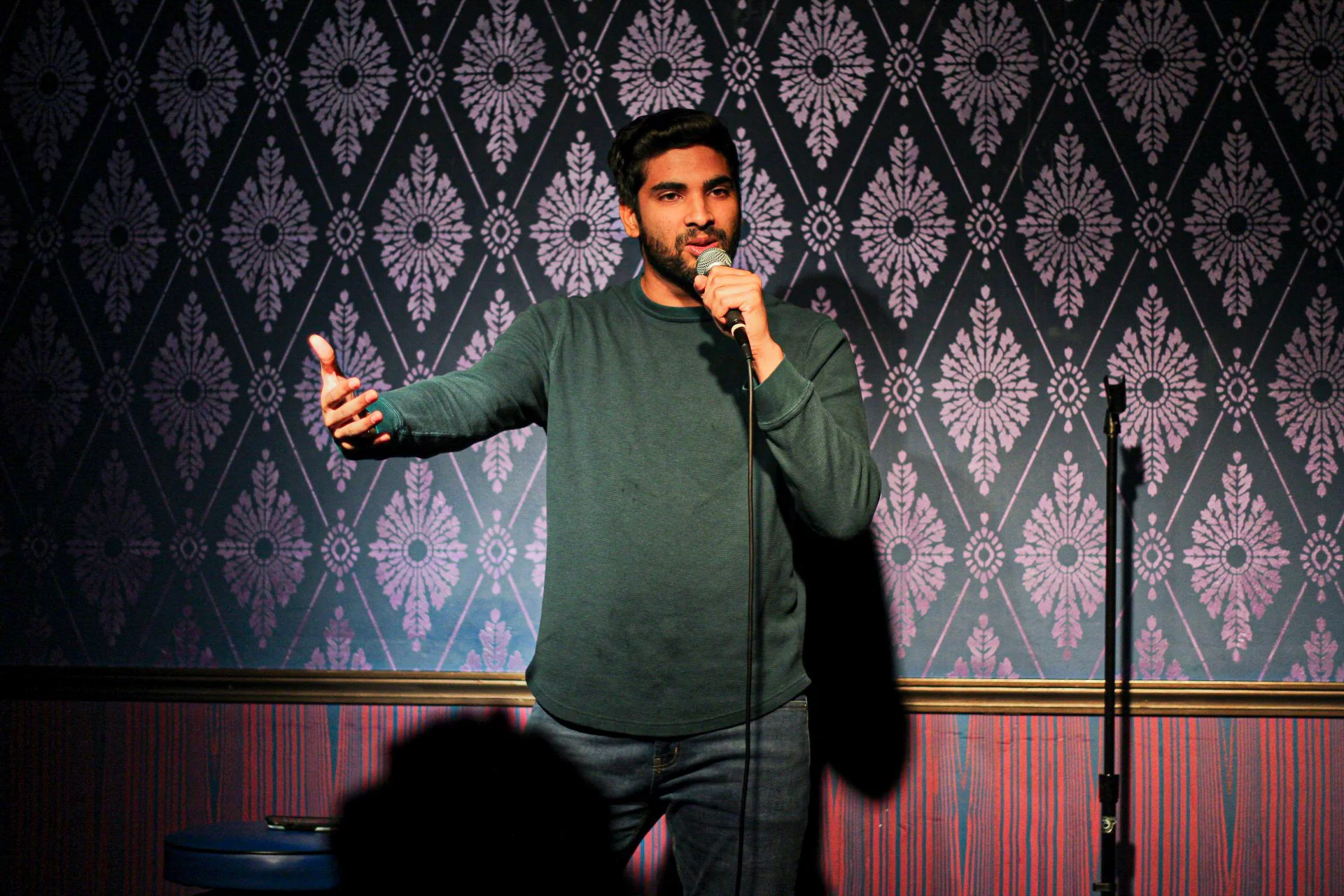 Comedian performing stand-up on stage while holding a microphone and gesturing to the audience.