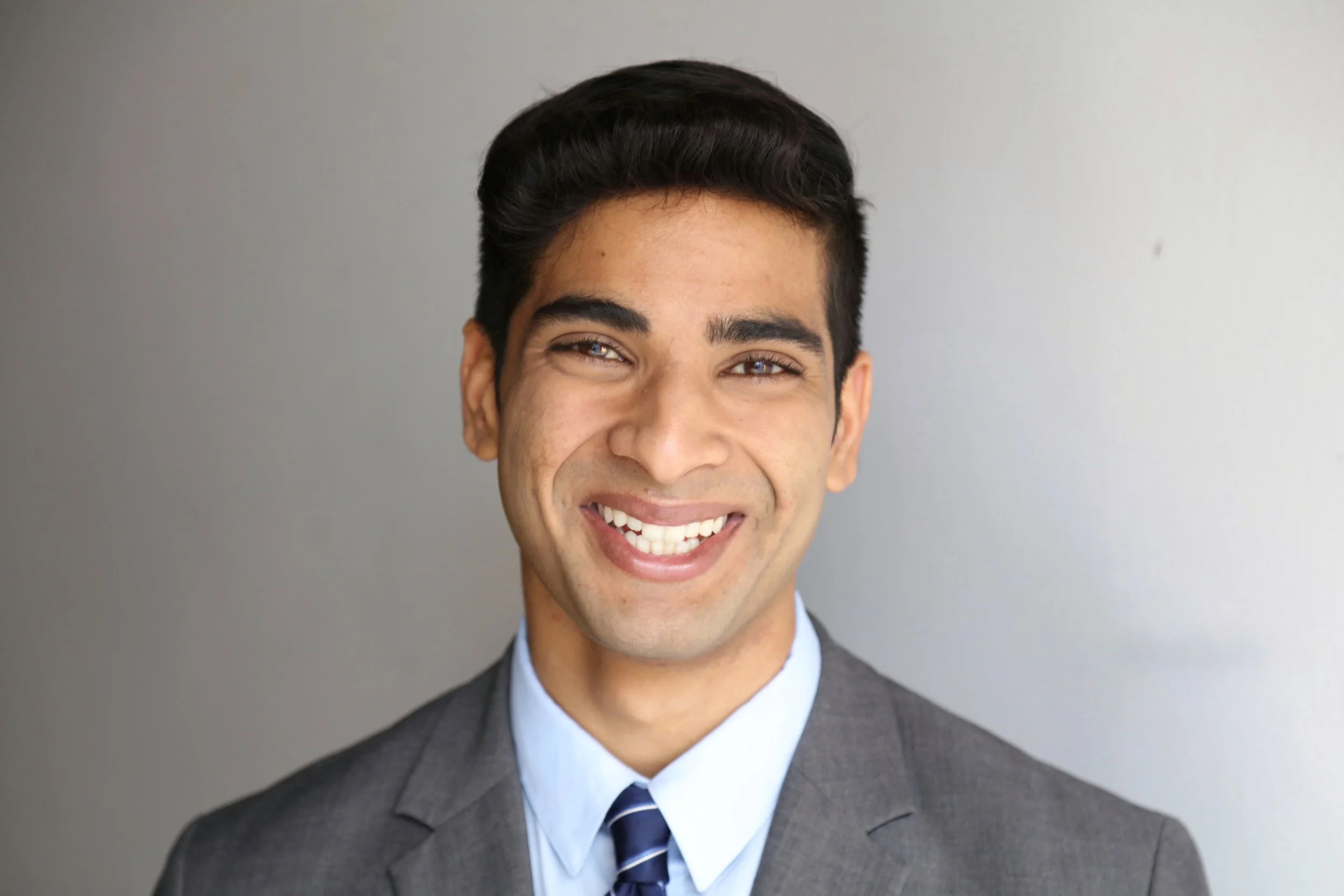 Smiling young man in a gray suit, white shirt, and striped tie against a light gray background