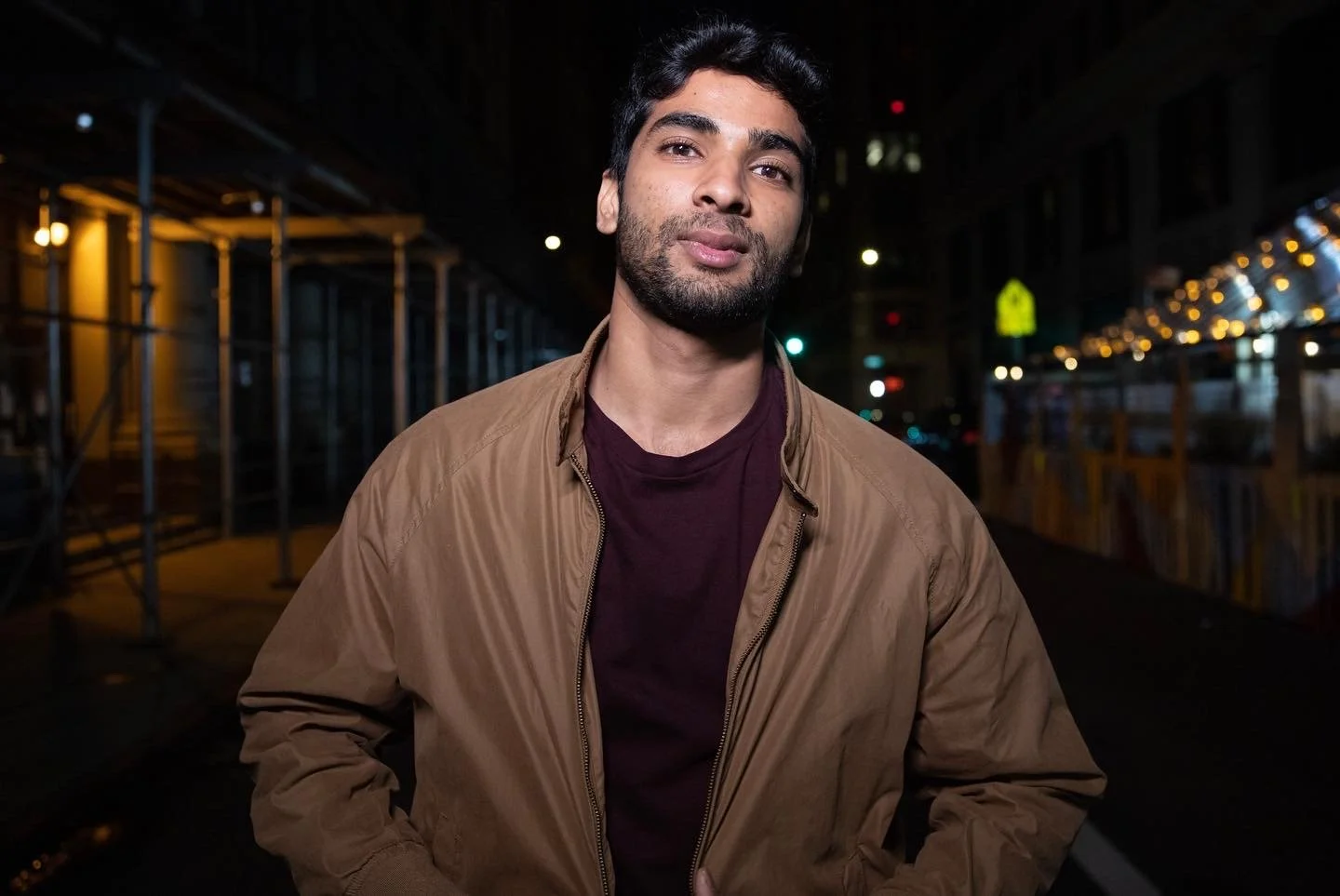 Man in a brown jacket standing outdoors at night on a city street with blurred lights
