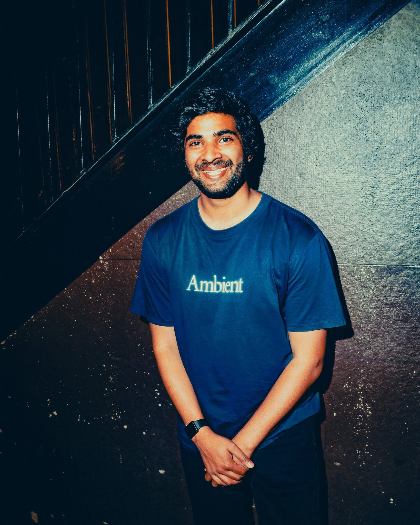 Smiling man with curly hair wearing a blue “Ambient” T-shirt, standing against a dark textured wall