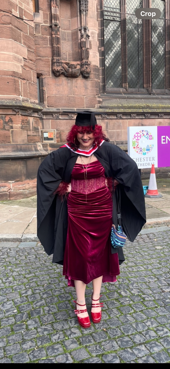 Woman with red curly hair wearing a graduation cap and gown, holding a purse, standing on cobblestone street outside of a historic building, smiling.