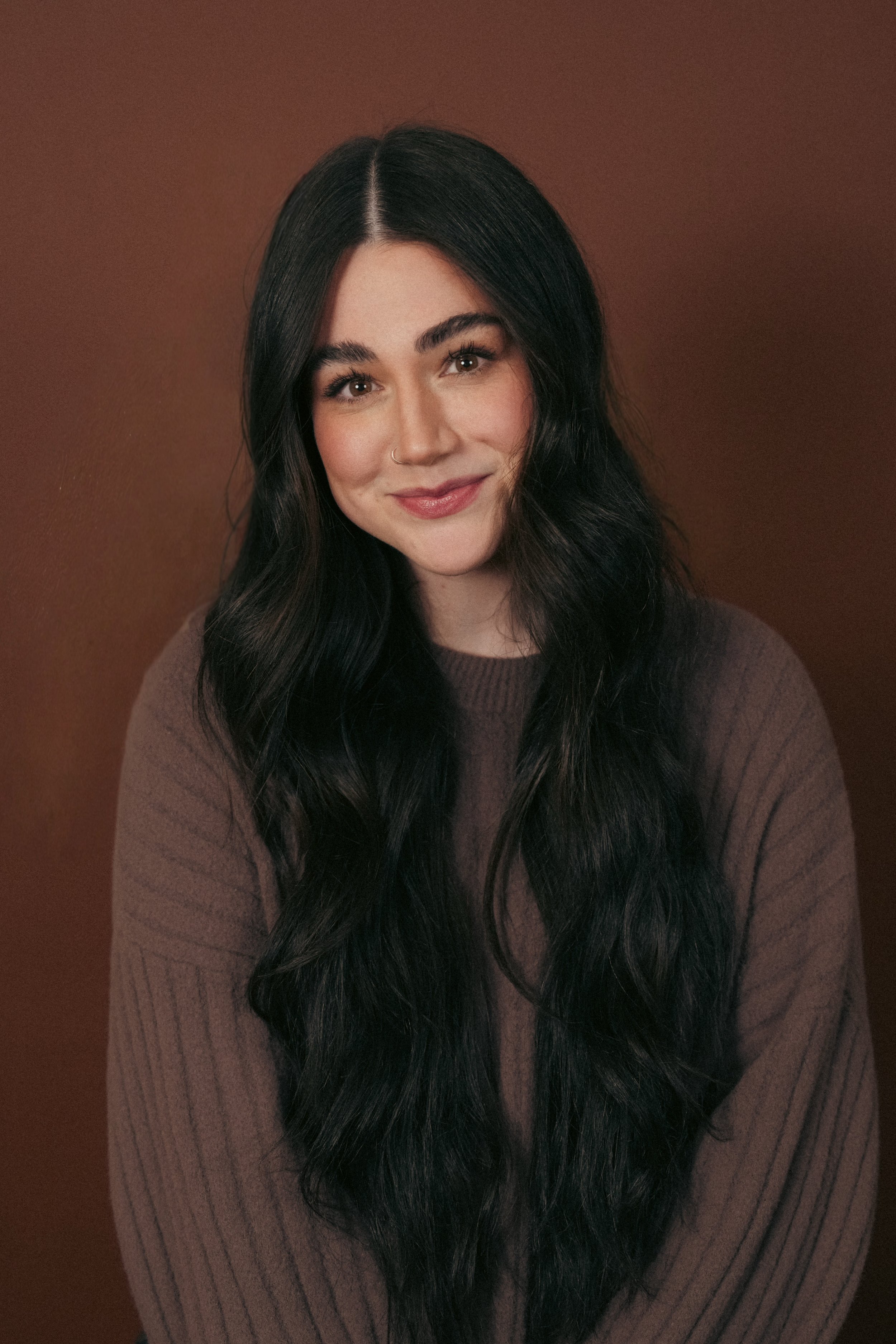 Portrait of a woman with long black hair, light skin, and a warm smile, wearing a brown knitted sweater, against a brown background.