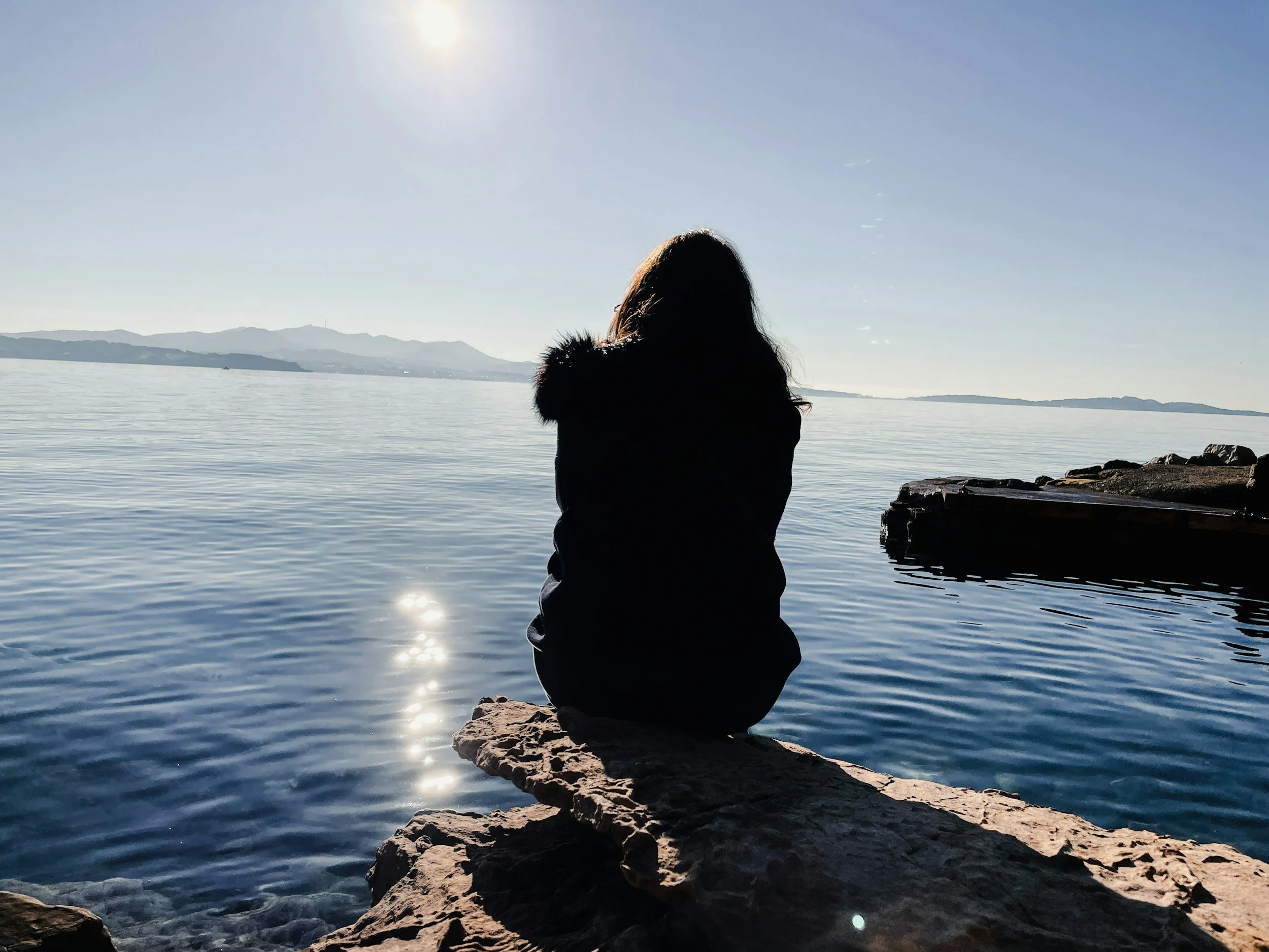 Looking at person from behind who is sitting on rock overlooking ocean with mountains in the background and sun high in the sky