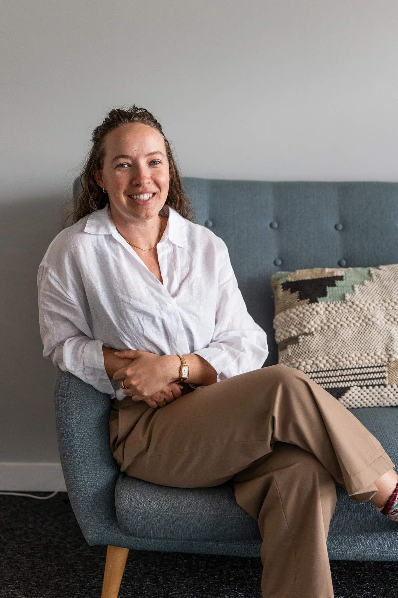 Smiling woman in a white blouse and tan pants sitting on a blue armchair beside a textured pillow