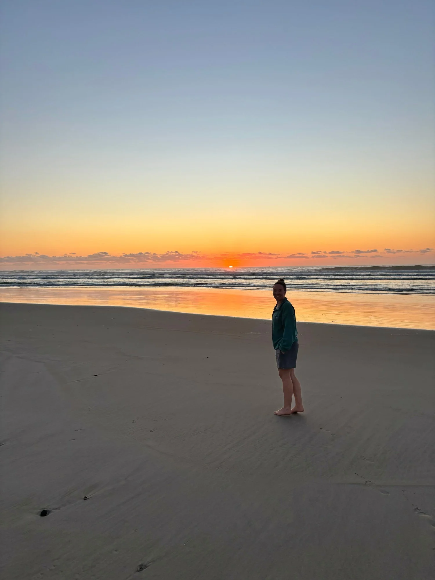 Woman standing barefoot on a sandy beach as the sun rises over ocean waves