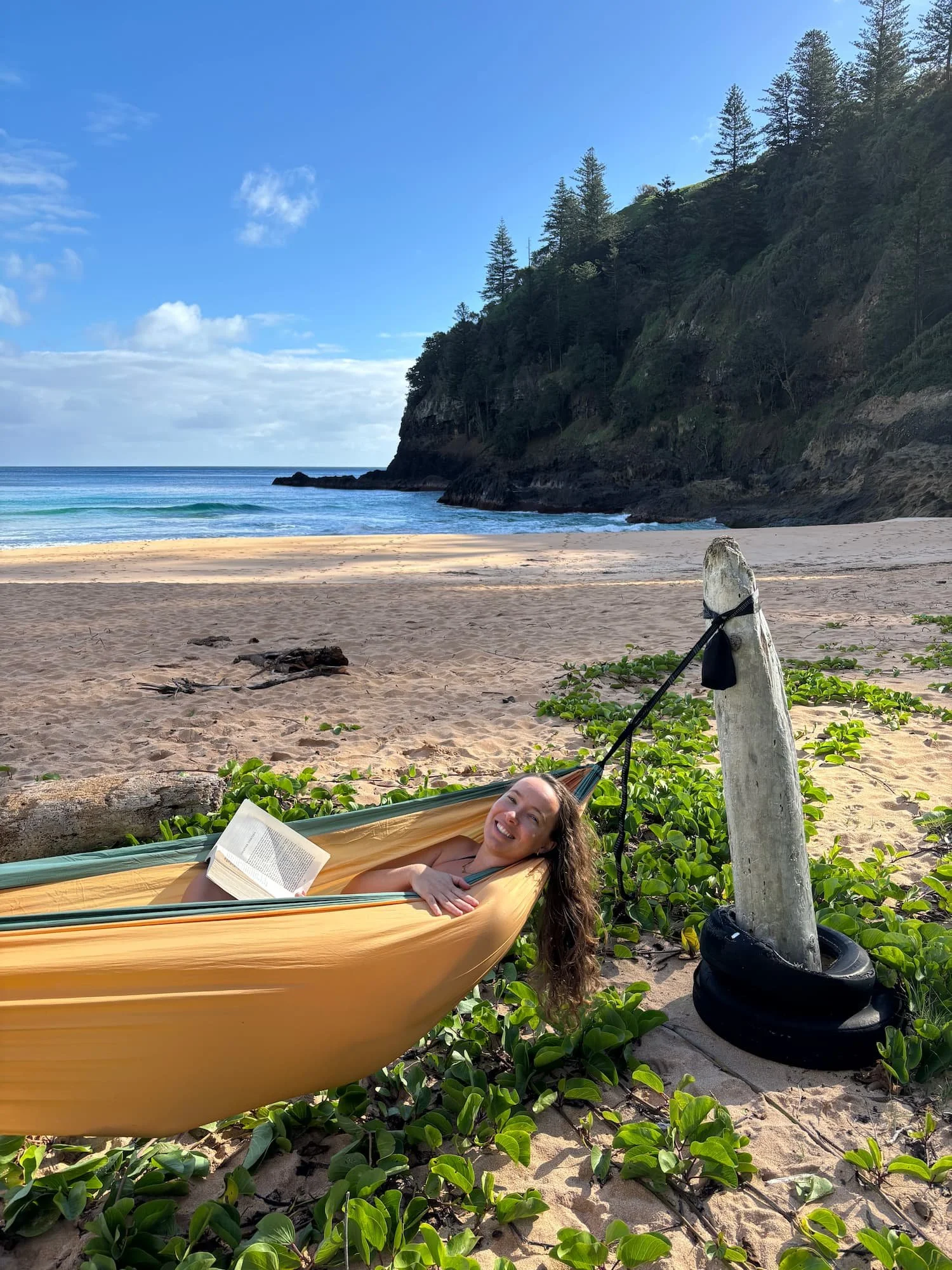 Woman reading a book in a hammock on a sandy beach beside turquoise ocean water and a tree-covered rocky cliff