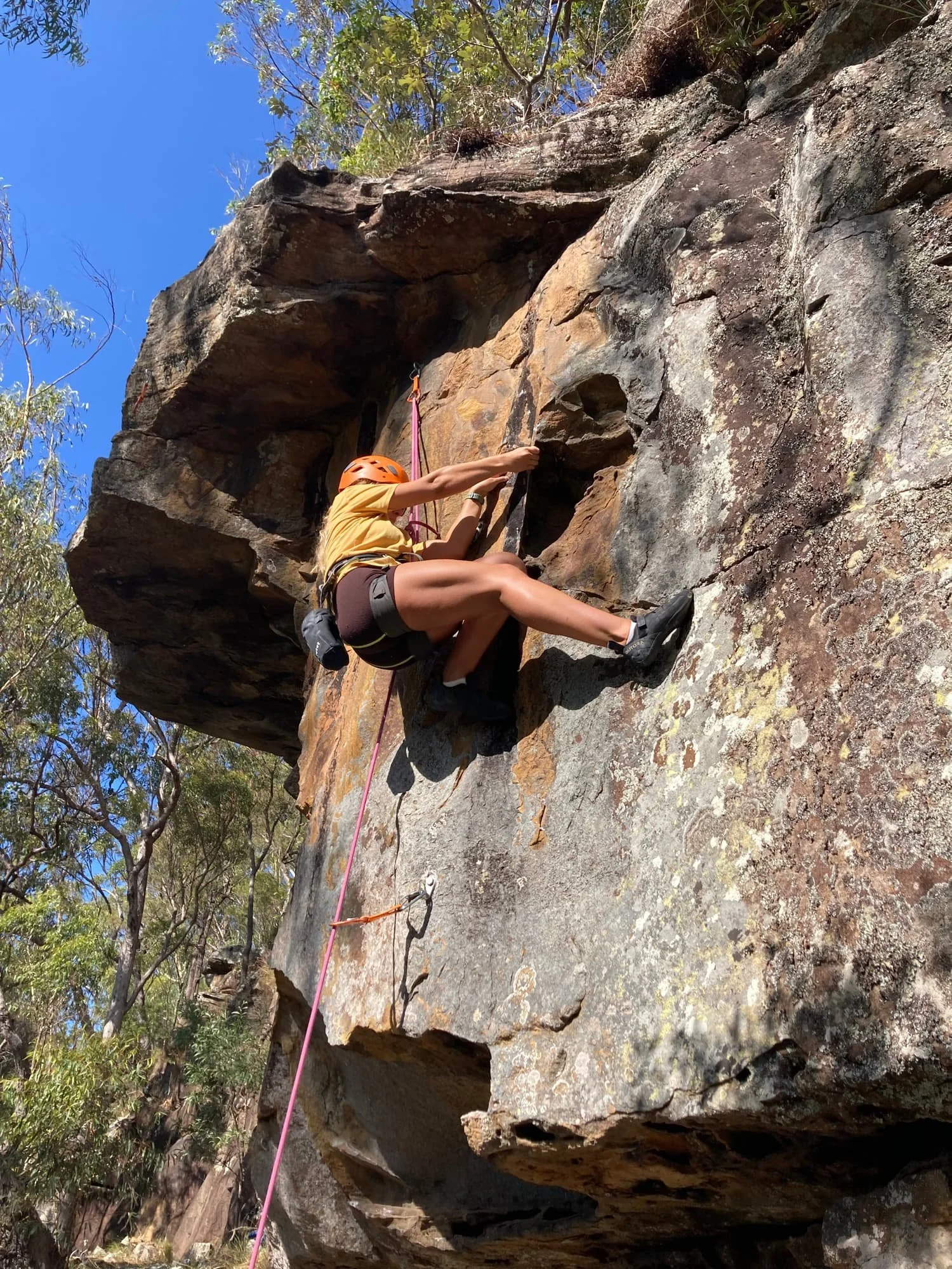 Rock climber wearing a helmet and harness ascends a sandstone cliff with a rope under a clear blue sky