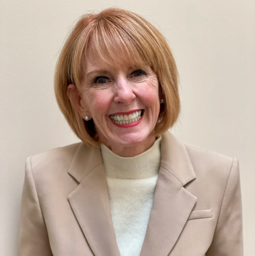 A woman with short red hair, smiling, wearing a beige blazer and a light-colored turtleneck, standing against a plain off-white wall.
