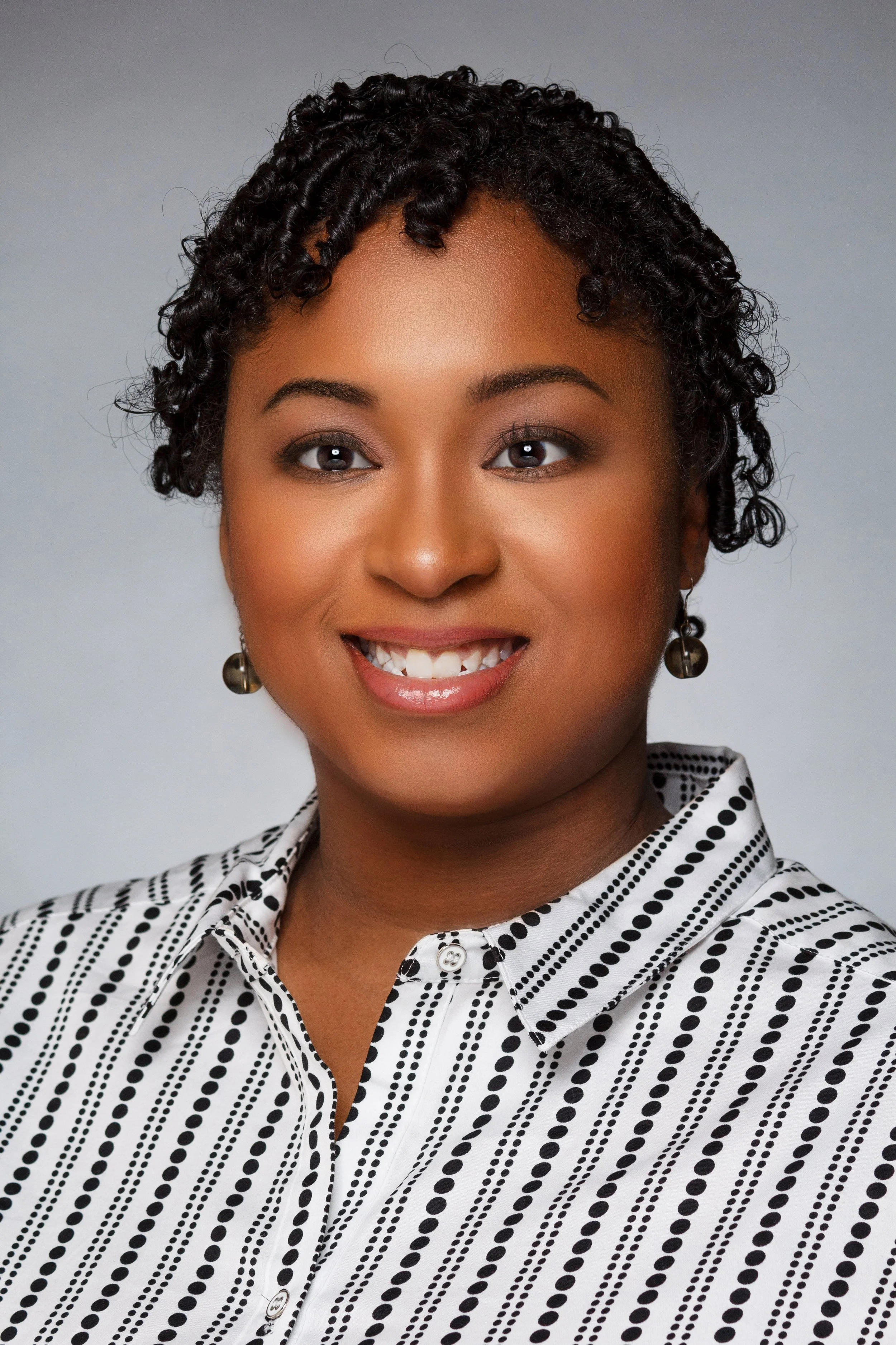 Close-up of an African American woman with short, tight curls, smiling and wearing earrings and a black-and-white polka dot blouse.